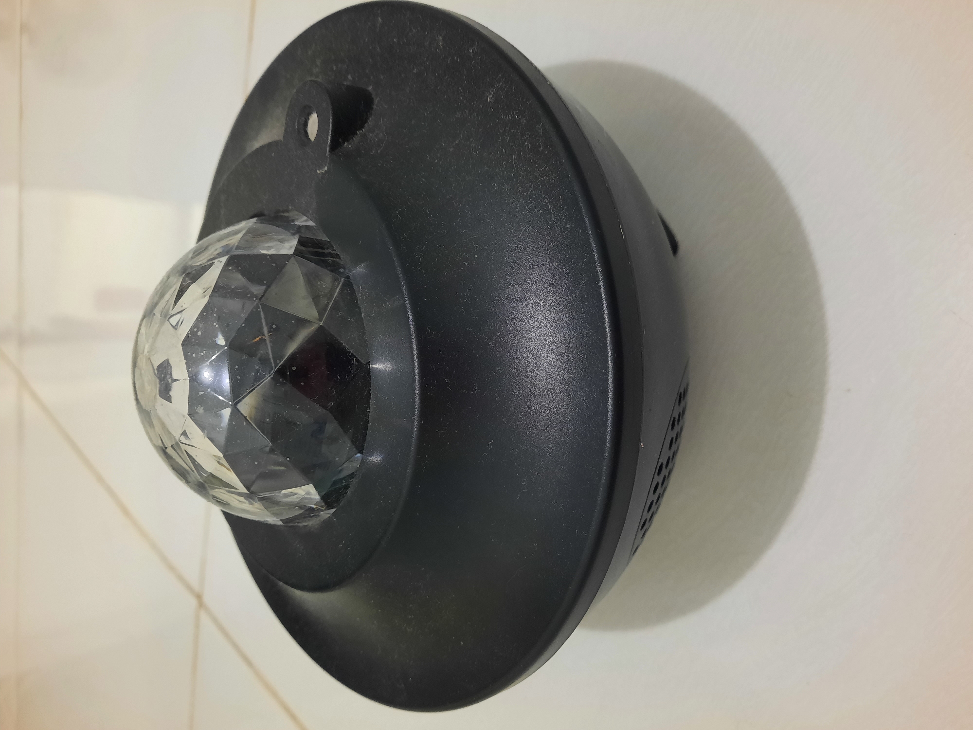 A black, round projector sits on a tiled floor. The projector has a faceted clear dome on top. The device appears to be a night light or mood light. The tiled floor is light-colored with visible grout lines. The lighting suggests an indoor environment. The image was taken in Lhokseumawe, Indonesia.