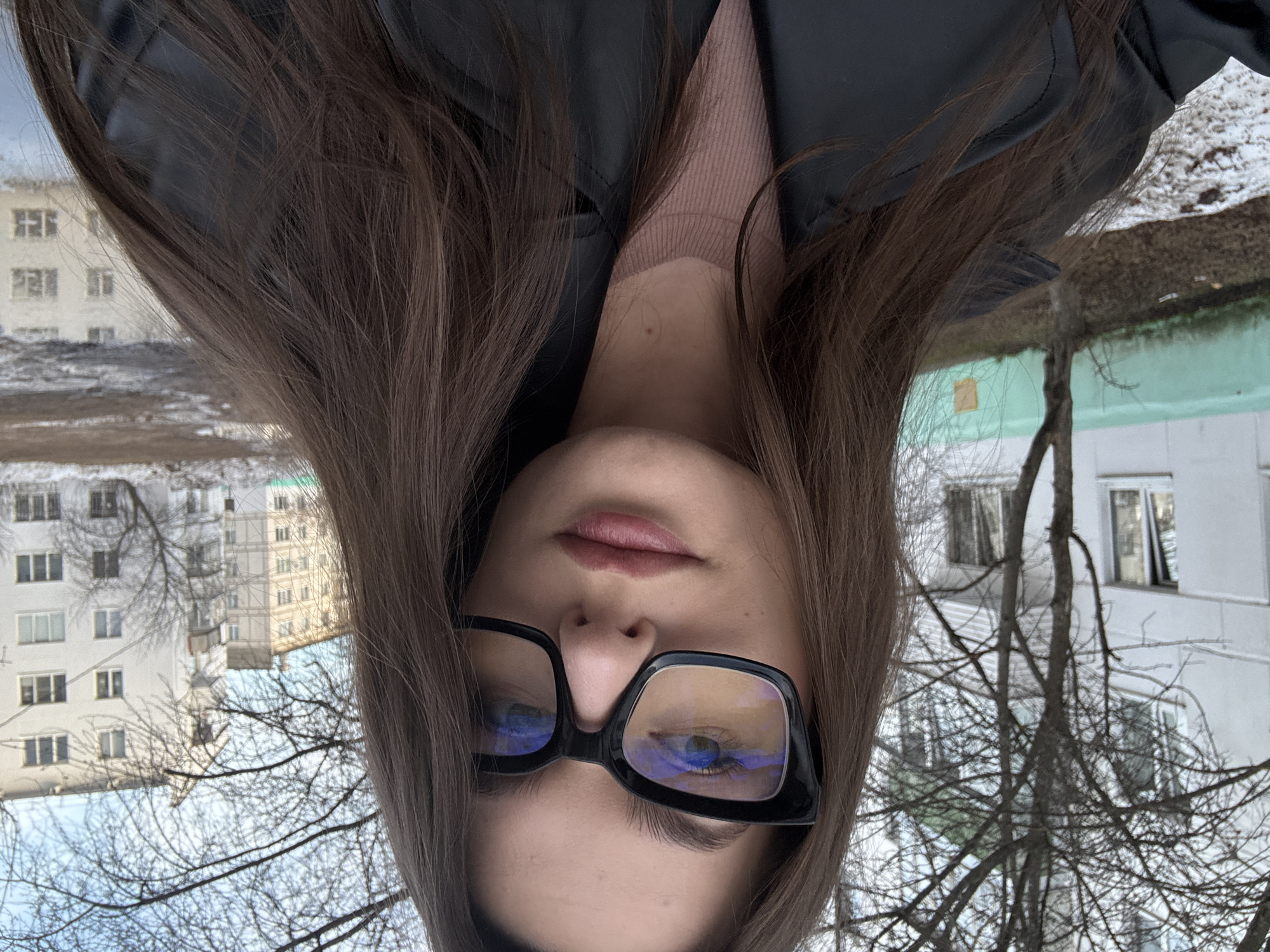 A young woman with long, straight dark brown hair, parted in the middle, is depicted in a close-up, likely self-portrait. She wears large, black-framed glasses with anti-reflective lenses showing blue and yellow reflections. Her lips are lightly colored, and her eyebrows are defined. She is dressed in a light brown ribbed crewneck garment beneath a black, leather-like jacket or coat. A small mole is visible on her upper chest/neck area.
The scene is an outdoor residential courtyard in the city of Chaikovsky, Russia. The ground displays patches of melting snow and exposed dark soil, alongside several bare deciduous trees with visible branches, suggesting a late winter or early spring season. In the background, multiple multi-story apartment buildings are present. A prominent white apartment block with numerous windows and balconies is visible to the left, partially obscured by trees. Further back and to the right, a lighter-colored apartment building of similar architectural style can be seen. A segment of a low, faded teal-colored wall or fence is located in the mid-ground on the left. A large puddle or shallow body of water in the lower right reflects the sky and background structures. The buildings exhibit architectural characteristics consistent with Soviet-era residential developments common in Russian urban environments.