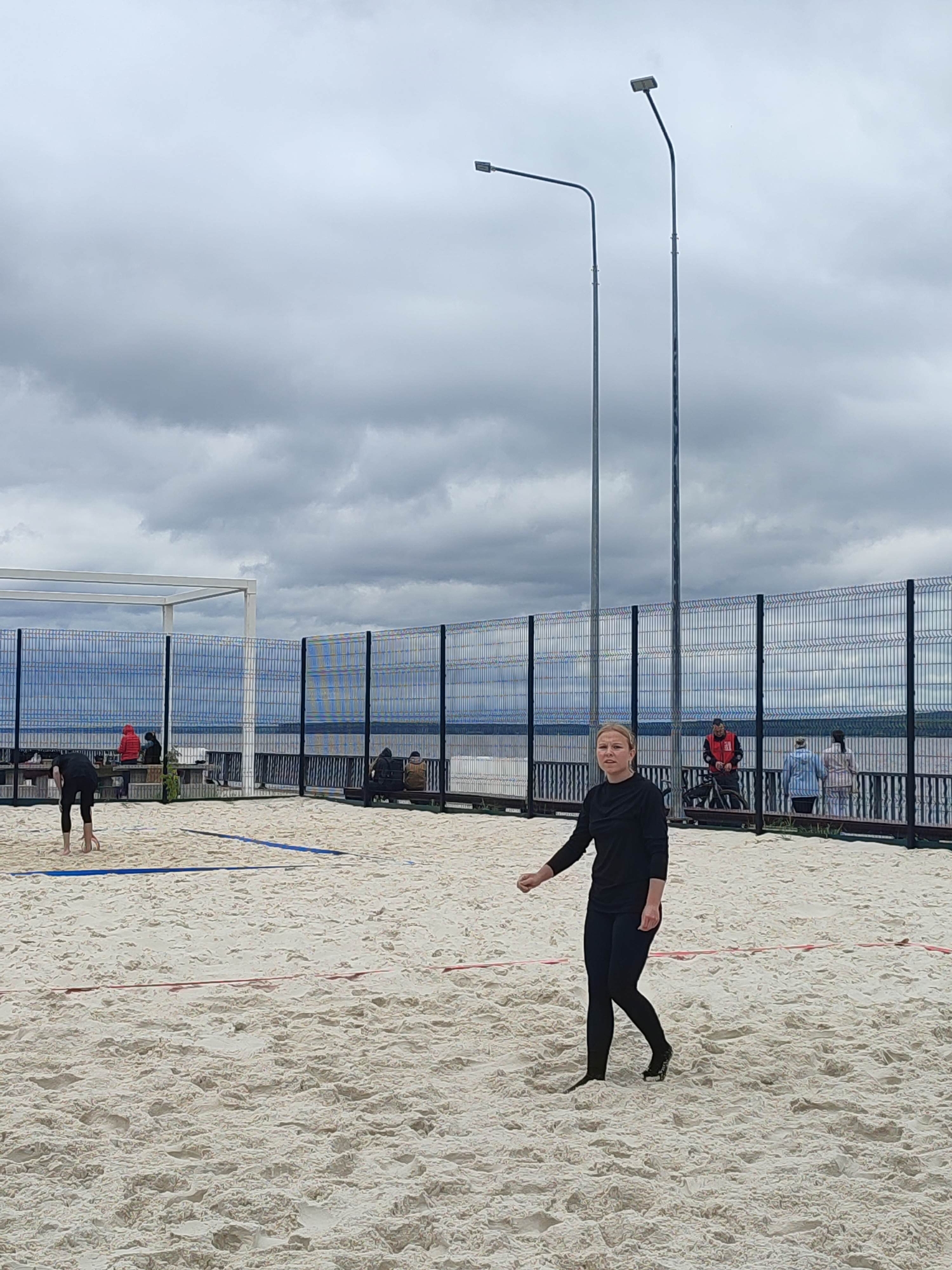 The image depicts a sandy beach volleyball court with a cloudy, overcast sky. Two streetlights with square lamps stand prominently in the background. In the foreground, a blonde woman wearing a black long-sleeved shirt and black leggings is in motion, appearing to be playing or practicing volleyball. Her body is angled, and her left hand is extended slightly forward.
Further back on the court, another person, dressed in black, is bending over, possibly retrieving a ball. Beyond the court, a black metal fence encloses the area. Behind the fence, a body of water is visible, suggesting a location near a lake or sea. Several other people are scattered in the background, some sitting on benches or standing near a wooden structure. One person in the background wears a bright red jacket, and another wears a blue and white patterned outfit.
The overall mood is subdued, likely due to the overcast weather. The sand is light in color, with visible footprints and indentations, indicating recent activity. The scene appears to be a public recreational area. There is no visible text in the image.