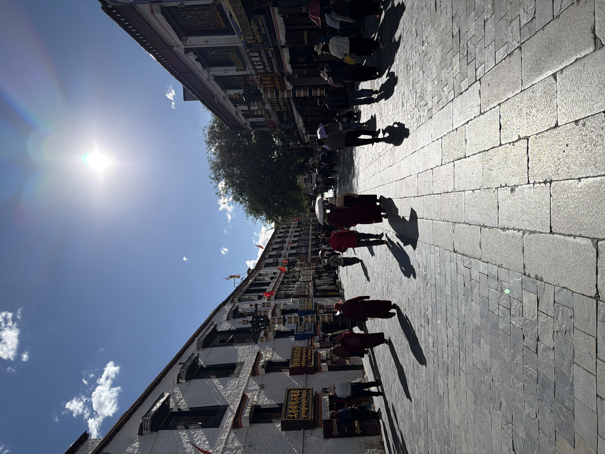 The image depicts a bustling, sunlit street scene in a Tibetan cultural area, identified as Chengguan District, China. The paved street, composed of various grey stone blocks, stretches into the distance, flanked by traditional multi-story buildings on both sides. A strong mid-day sun, visible in the upper left with a bright lens flare, creates long, distinct shadows of pedestrians extending across the pavement.
Numerous people are seen walking along the street. Several individuals are dressed in traditional maroon or red robes, indicative of monks or pilgrims, while others wear more casual clothing. Some carry umbrellas for sun protection. A large, green-leafed tree stands prominently on the left side, providing a natural element amidst the architecture.
The buildings exhibit characteristic Tibetan architectural styles, featuring white-washed walls, dark window frames, and intricate decorative elements. Small red flags hang from the building on the left. Shopfronts display signs in both Chinese and Tibetan script. Visible texts include "土登卓嘎木碗店" (Tudeng Zhuoga Wooden Bowl Shop), "店" (Shop), "吉祥" (Auspicious), and "ཕྱིང་དཀར་ཞྭམོ་ཆེད་འཚོང་ཁང་།" (White Felt Hat Special Sales Store). The sky is a clear, bright blue with a few scattered white clouds. The scene conveys a lively commercial or cultural district under brilliant sunshine.