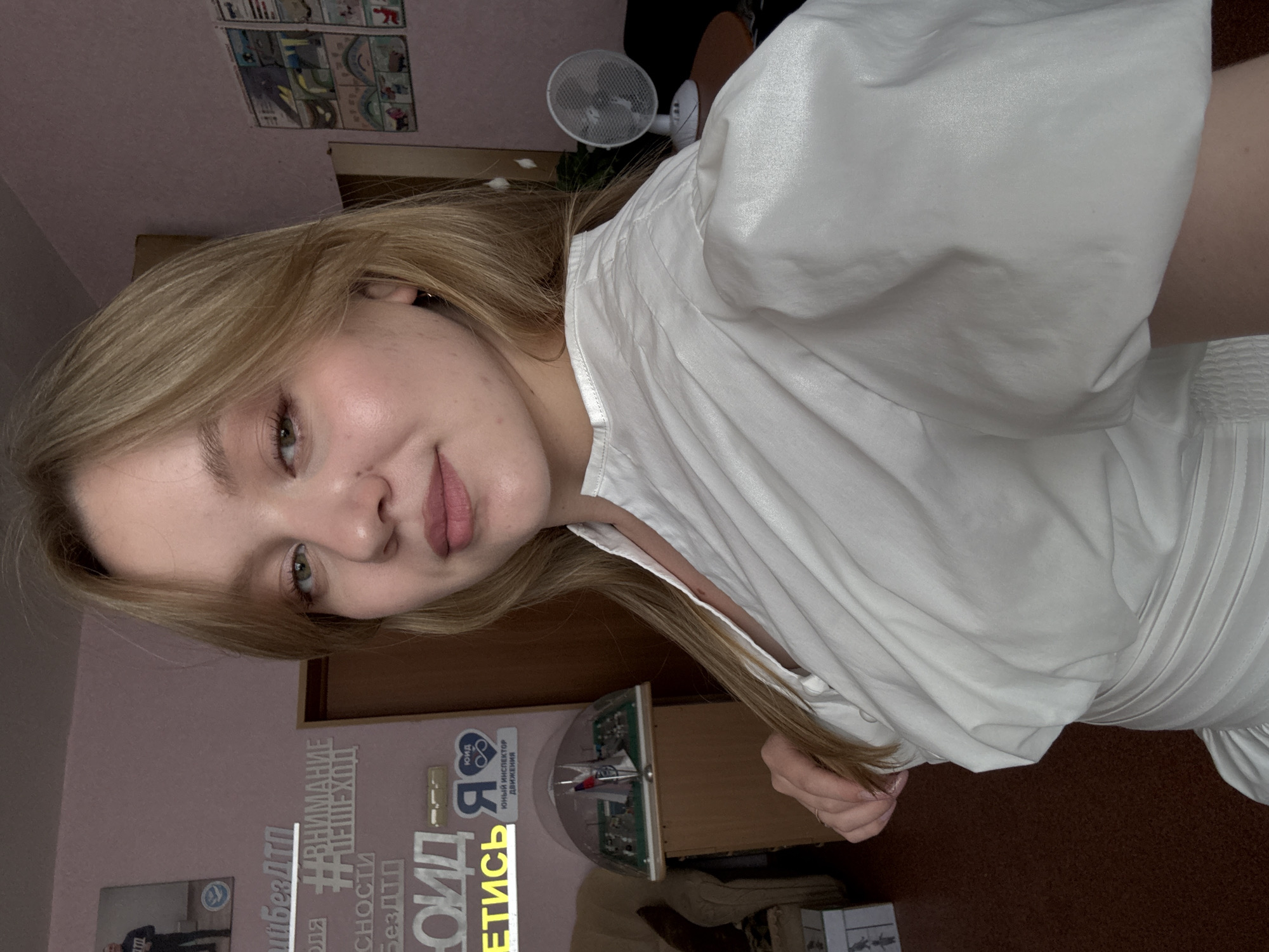 A young woman with blonde hair, wearing a white dress, is pictured in a selfie. The background shows a pink wall adorned with several posters and signs with text in Russian, indicating a location in Chaikovsky, Russia. One poster features a person in uniform, while others display slogans and graphics. To the left of the woman, a display of miniature buildings is visible on a table. A small white fan is placed on a table to the right. The overall setting appears to be an indoor space, possibly a room or office, in Chaikovsky, Russia.