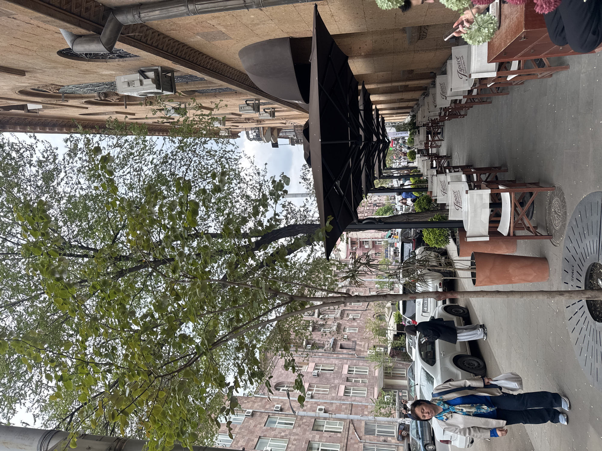 This daytime image captures a bustling street scene in Yerevan, Armenia, characterized by a mix of historical architecture and modern urban life under an overcast sky.
The foreground is dominated by a large tree with vibrant green foliage, partially obscuring the view of the street and buildings beyond. To the right of the tree, a sidewalk cafe stretches along the building, featuring rows of unoccupied wooden tables and chairs shaded by large, black retractable awnings. The white chair backs prominently display the text "J.space creative urban hub."
Along the paved sidewalk, several individuals are visible. In the immediate foreground, a person wearing a light-colored jacket, blue top, and dark pants stands, holding a white bag. Further down, another person dressed in a black jacket and grey sweatpants is engrossed in their phone, standing near a parked white car. Other figures are faintly visible in the distance. Parked cars line the street, one with a visible Armenian license plate displaying "13 AV 31."
The backdrop consists of multi-story buildings constructed from a distinctive pink-toned stone, typical of Yerevan's architecture. These buildings feature numerous windows, some arched, and multiple air conditioning units attached to their facades. Architectural details, including cornices and decorative elements, are discernable on the building's upper sections. Large planters are placed along the sidewalk, contributing to the green urban environment.