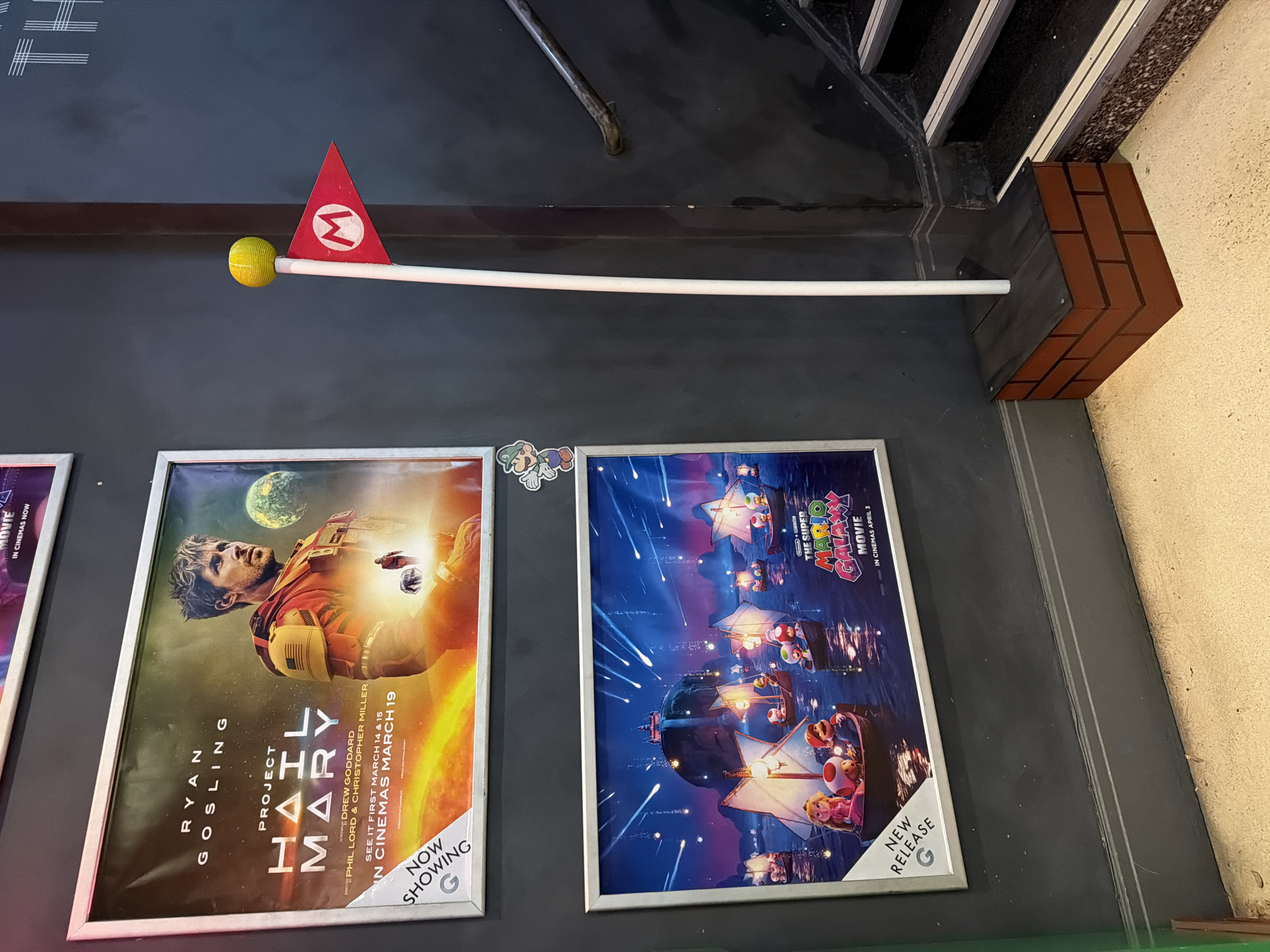 The image shows two movie posters and a Mario flagpole positioned on a wall in an unidentified indoor location in London, United Kingdom.
To the left, a movie poster features Ryan Gosling in the film "Project Hail Mary," showing the "Now Showing" banner at the bottom right, and is listed with a "G" rating. To the right of it, a second movie poster displays "The Super Mario Galaxy Movie," marked as a "New Release" with a "G" rating and "In Cinemas April 3" written in the text.
Above these posters, a white flagpole extends horizontally. At one end is a yellow ball, and the other end features a red flag with the letter "M" in a white circle. The flagpole appears to be held in place by a decorative block with a brick pattern.