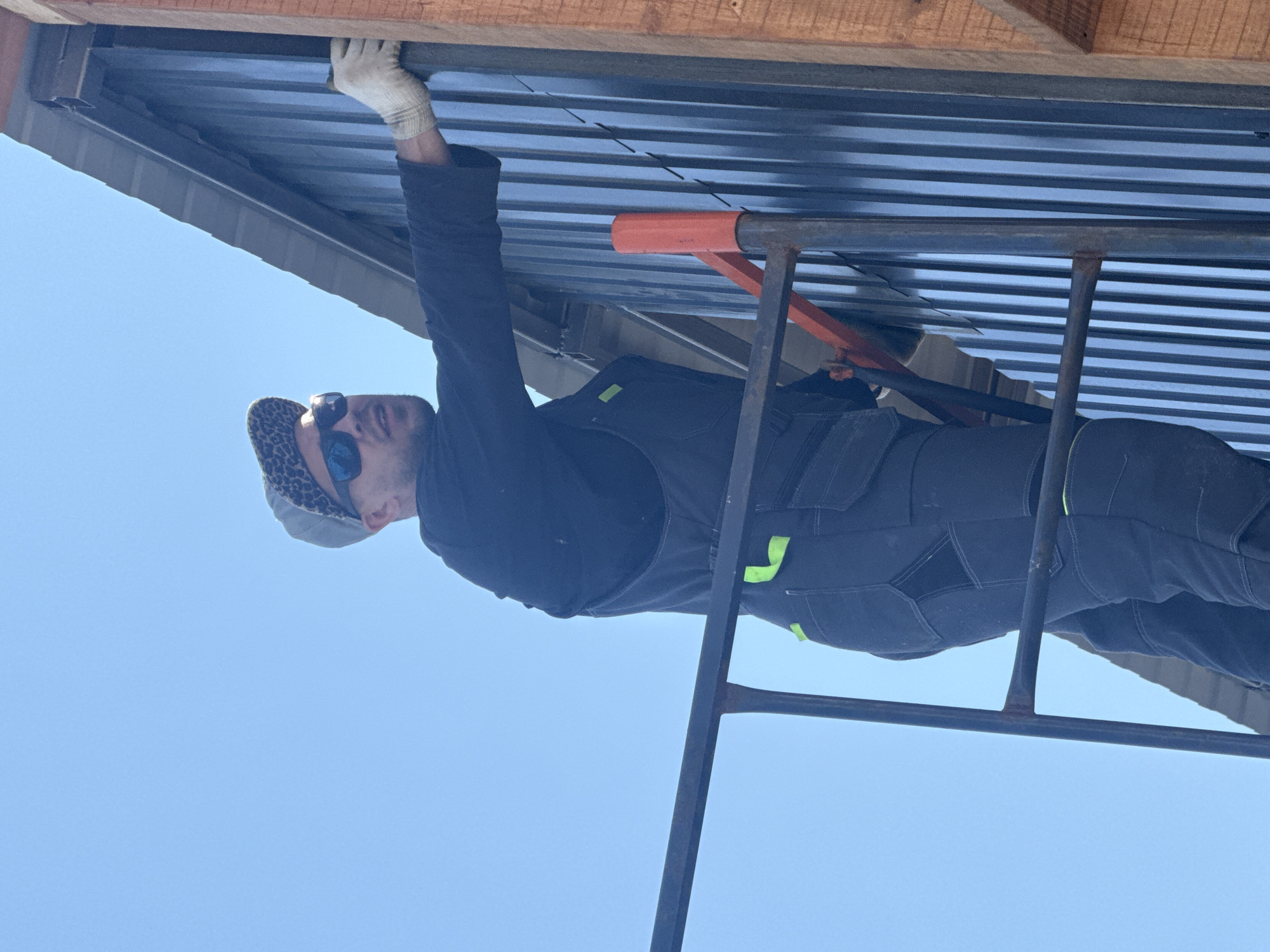A man wearing a hat, sunglasses, and work clothes is working on a corrugated metal roof. He is wearing gloves on his left hand and standing on a metal scaffold. The sky is blue. The location is Unknown, Unknown.