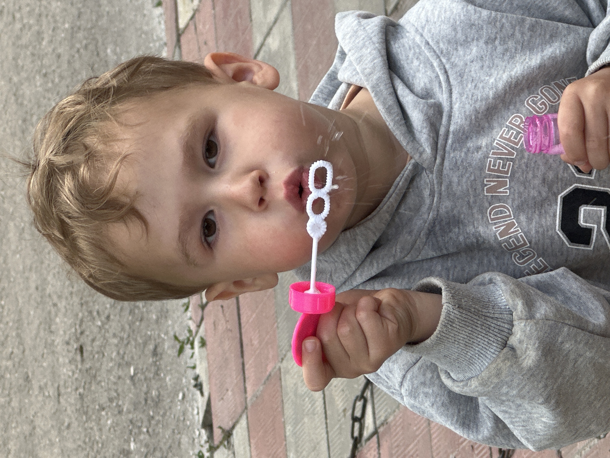 The image shows a close-up of a young child blowing bubbles. The child has light brown, wavy hair and brown eyes. They are wearing a gray hooded sweatshirt with white text that reads "LEGEND NEVER GON". The child is holding a pink bubble wand with a white ring, from which a bubble is forming. The child's lips are pursed as they blow. The background appears to be an outdoor setting with a paved ground made of light-colored bricks and gray concrete. The lighting suggests it might be daytime, possibly overcast. The child's expression is focused on the activity of blowing bubbles. The location is identified as Nizhny Novgorod, Russia.