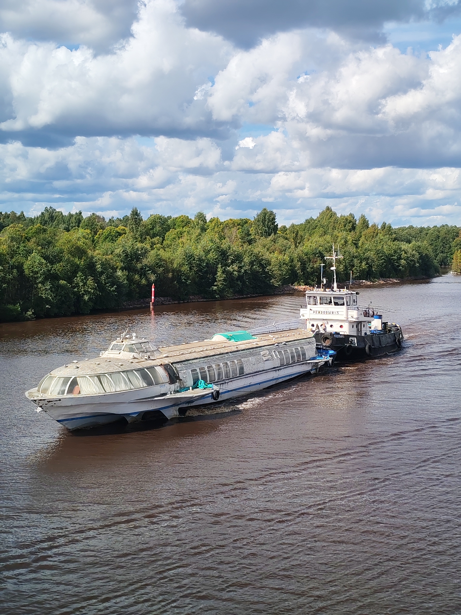 In Annensky Most, Russia, a fleet of two vessels navigates a wide, murky brown river under a dynamic sky filled with large, white and gray cumulus clouds. The primary vessel, a sleek, white hydrofoil with numerous large windows, is positioned in the foreground and moves from left to right. Its design suggests passenger transport capabilities, with white curtains visible behind the glass. Attached to its stern, a black and white tugboat, labeled "РЕЗДОВЫЙ-5," provides propulsion. The tugboat has a multi-level superstructure with windows and an antenna mast. The riverbanks are lined with a dense, lush green forest, with the trees displaying a variety of shades indicating their autumnal transition. A red and white buoy is visible in the river, marking a navigation channel or hazard. The water's surface is disturbed by the movement of the vessels, creating visible wakes and ripples. The overall scene captures a moment of transit on a river in a natural, forested environment.