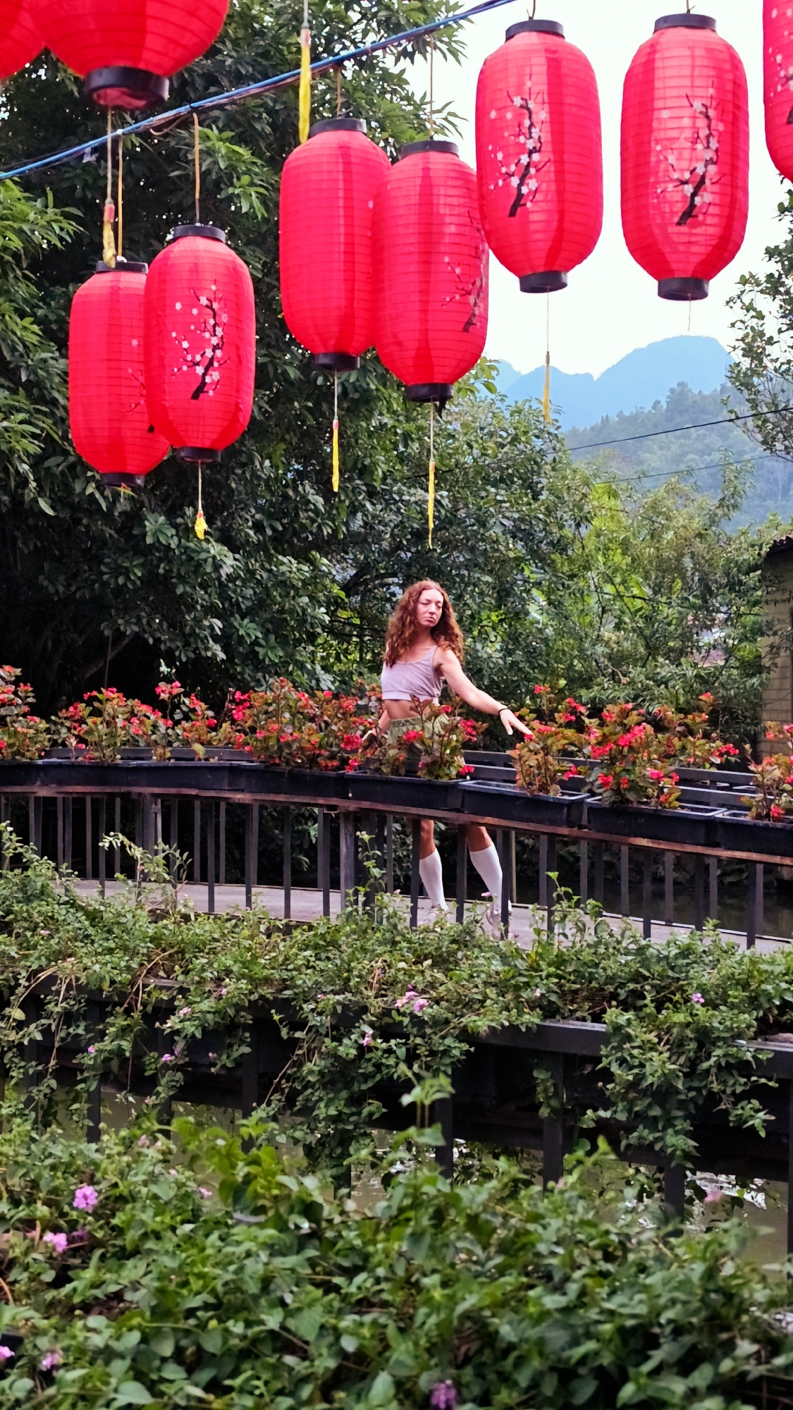 A full shot captures a fair-skinned woman with curly red hair, wearing a light pink top and dark green shorts, standing on a bridge decorated with flower boxes filled with red and green plants. The woman is wearing white, calf-length socks and her right hand is extended towards the plants. Above the bridge, several red Chinese lanterns hang, suspended from an unseen structure. Green trees and foliage surround the bridge. Distant, sloping mountains are visible in the background. The image appears to be taken in Yên Minh Commune, Vietnam.