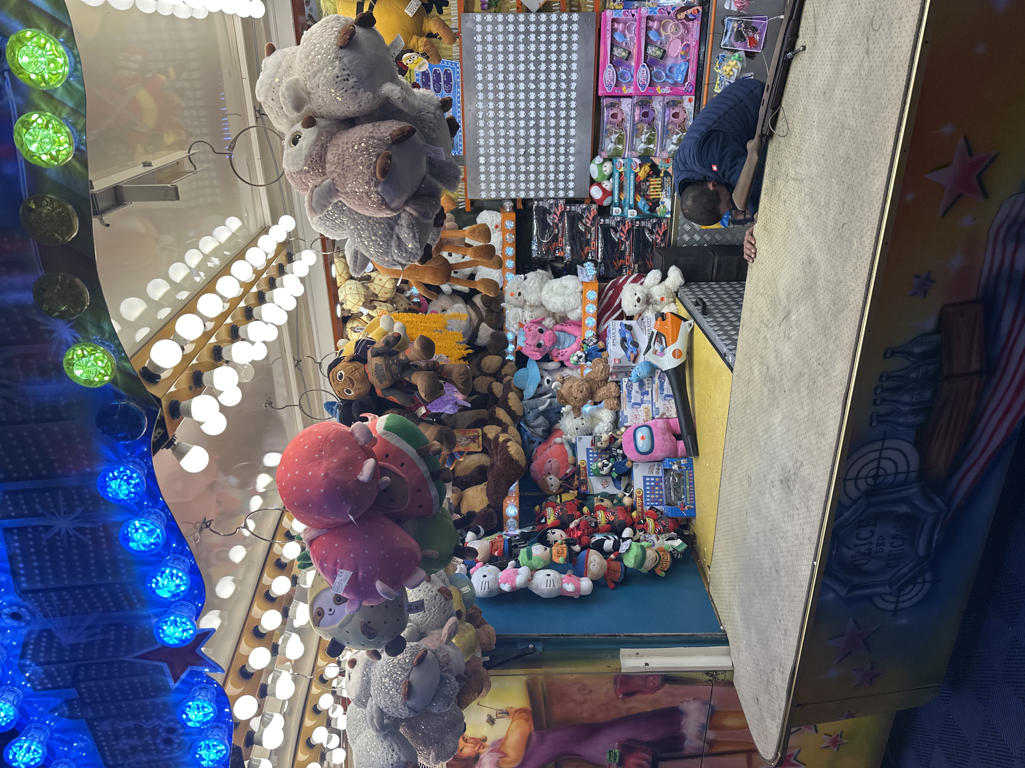 The image depicts a carnival game booth in Bruchhausen-Vilsen, Germany, likely at the "Bruchhäuser Markt" fair. The booth is filled with an assortment of plush toys hanging from above, including various animals like sheep and bears, as well as character plushies. Below the toys are shelves stocked with various items, including toy cars, dollhouses, and other games. A man, wearing a dark blue jacket, is positioned behind the counter, leaning forward and appearing to be preparing a rifle, possibly for a shooting game. The booth is brightly lit with strings of blue and white lights overhead, creating a festive atmosphere. The counter itself is covered with a textured material, and the side of the booth is decorated with painted targets and lettering that appears to read "Polizei Dep. Chicago," suggesting a police or shooting-themed game.