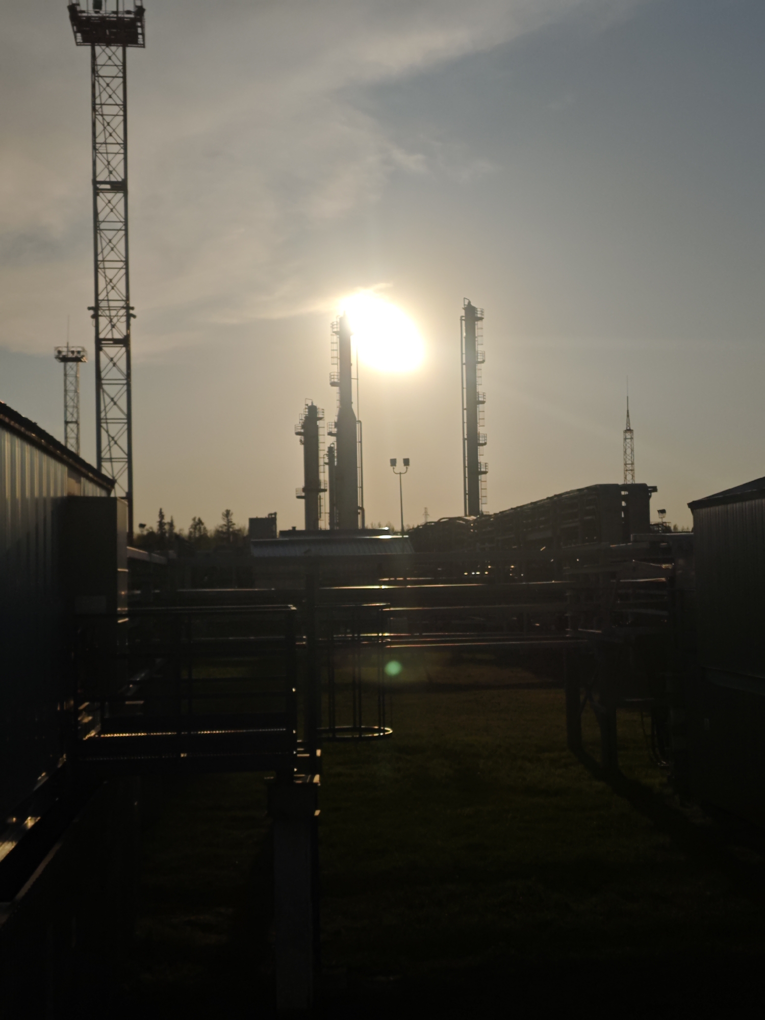 The image depicts an industrial landscape at sunset in an undetermined location in Russia. Several tall metallic towers and chimneys rise above the horizon, silhouetted against the bright sunlight and sky. Pipes and support structures traverse the foreground, with patches of vegetation visible beneath. Buildings flank the sides of the frame. The lighting is harsh, creating deep shadows and obscuring details in the lower portion of the image. There are no people present in the image.
