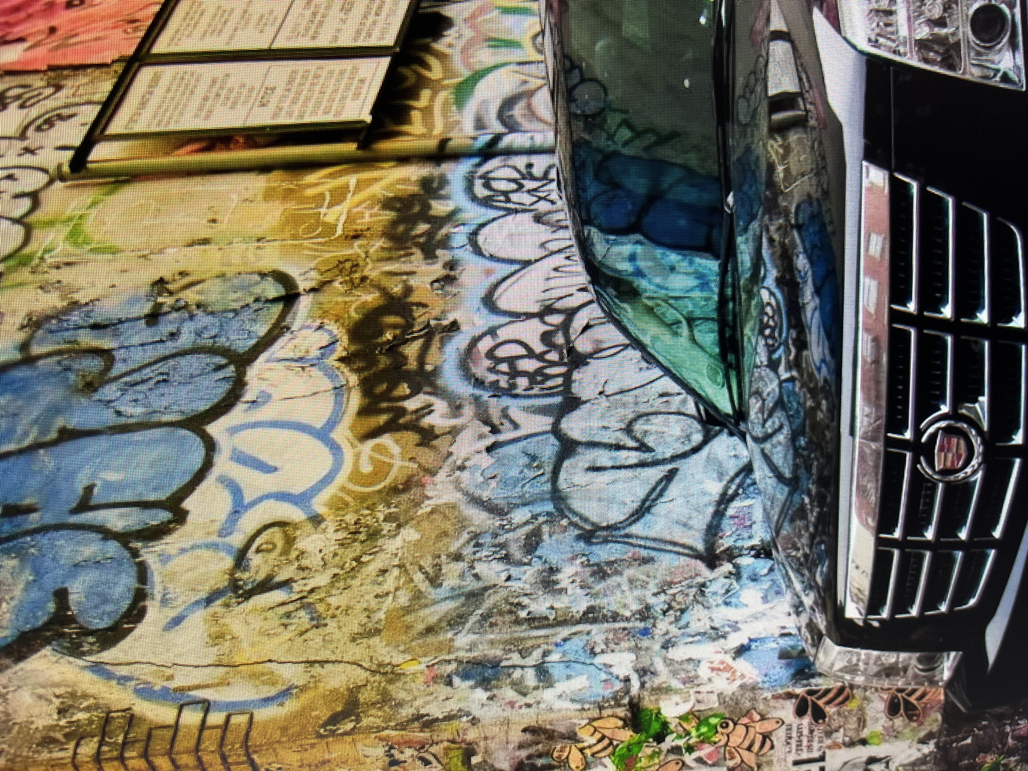 An outdoor scene depicts a black SUV parked in front of a graffiti-covered wall. The wall is extensively decorated with colorful spray-painted tags and murals, predominantly in shades of blue, yellow, and pink. Some of the graffiti appears to be in a bubble letter style, while other sections feature more abstract designs. A thin, vertical metal pole is visible on the right side of the wall, with two framed notices attached to it. The front grille of the SUV is prominent in the lower portion of the image, featuring horizontal chrome bars and the Cadillac emblem. The vehicle's hood and windshield reflect the graffiti-covered wall. The environment appears urban, with the wall suggesting an alleyway or a side street. The overall impression is one of urban decay and artistic expression.