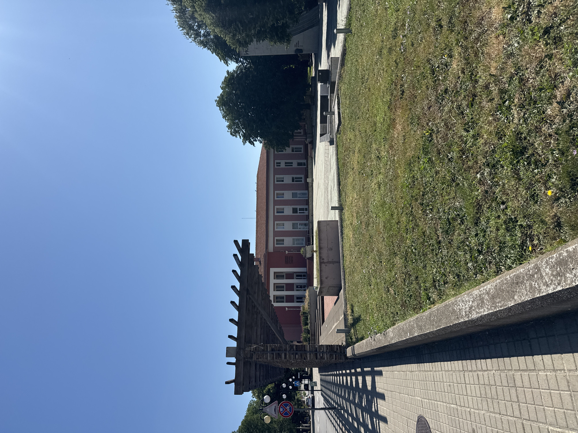 The image depicts an outdoor scene in the city of Shabla, Bulgaria, on a clear, sunny day. In the foreground, a paved walkway with a checkered pattern is visible on the left, casting a geometric shadow from a wooden pergola structure. A concrete border separates the walkway from a grassy area to the right. The pergola itself is constructed with weathered wooden slats supported by stone pillars.
In the middle ground, a red building with a tiled roof and numerous windows dominates the scene. The building has a neoclassical architectural style with prominent white columns supporting an overhang. In front of the building, there are some low-lying bushes and a paved area. To the right of the red building, a large, dark green tree stands tall, and further to the right, a tall, light-colored monument or sculpture is partially visible.
The background is a clear, bright blue sky. Traffic signs are visible on the left side of the walkway, including a traffic light and a circular no-entry sign. A white car is also seen on the road to the left. The overall environment suggests a public space, possibly a town square or a park area adjacent to a civic building.