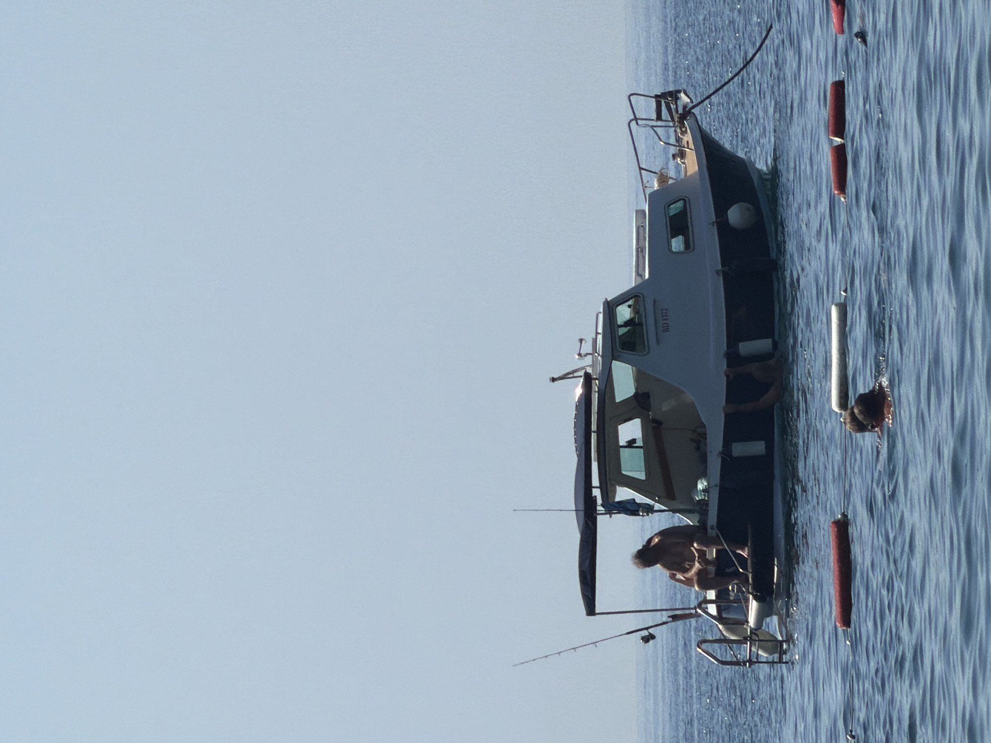 A boat is moored in the water off the coast of Budva, Montenegro. The boat has a blue hull and a white cabin with windows. A man is sitting on the edge of the boat, facing away from the camera. There are two fishing rods attached to the boat. In the water, two people are swimming, and several buoys are floating on the surface. The sky is clear and blue.