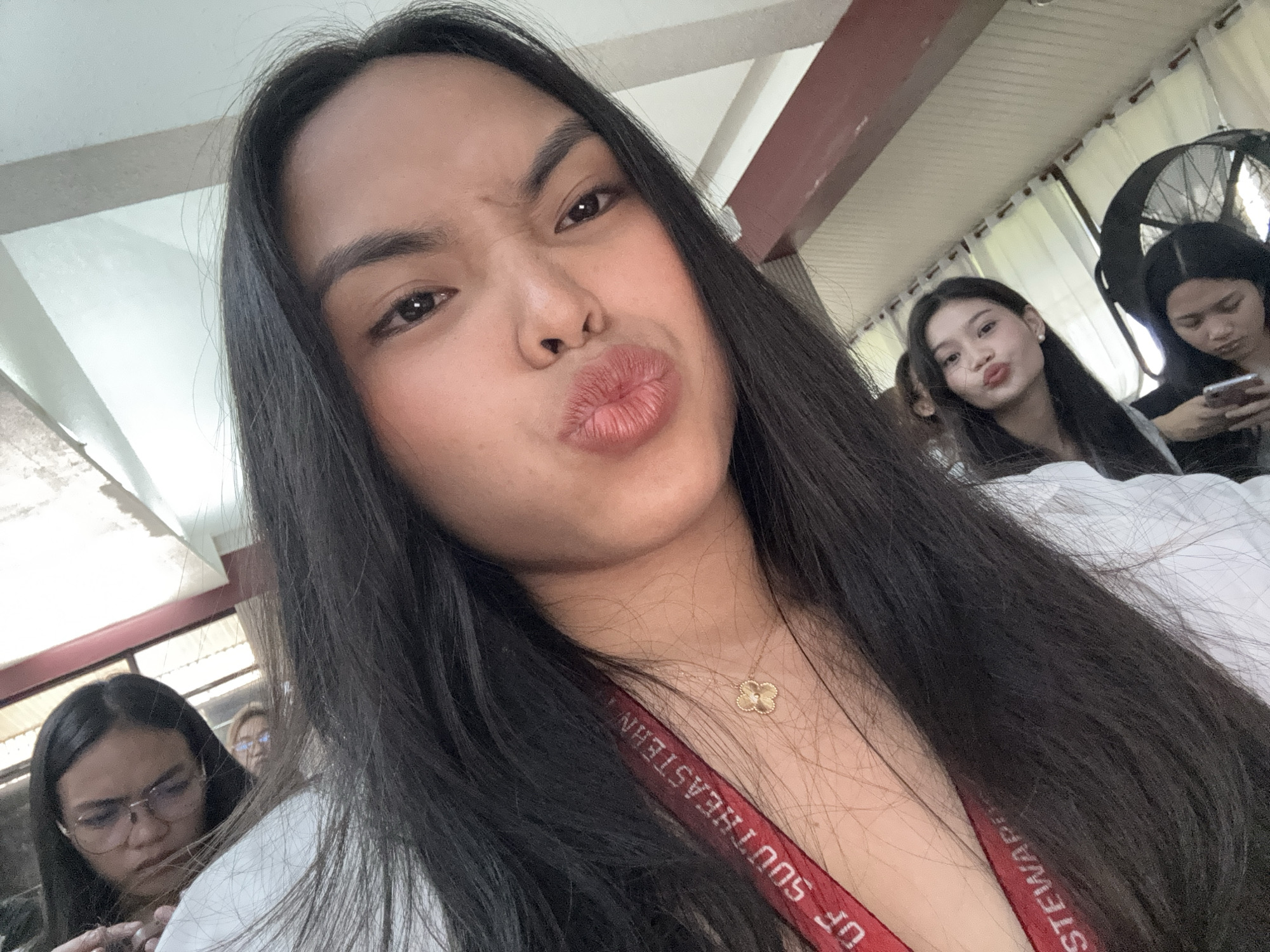 The image is a close-up selfie of a young woman with long, dark hair, making a playful face with pursed lips. She is wearing a white top and a red lanyard with text that appears to be "UF SOUTHEASTERN". In the background, several other young women are visible, some looking at their phones or down, and one is also making a similar facial expression. The environment appears to be indoors, possibly a classroom or common area, with light-colored walls and ceiling. Based on the text on the lanyard, the location is likely the University of Southeastern Philippines, which has campuses in Davao City, Philippines. There are no other visible details to further specify the exact location within Davao City.