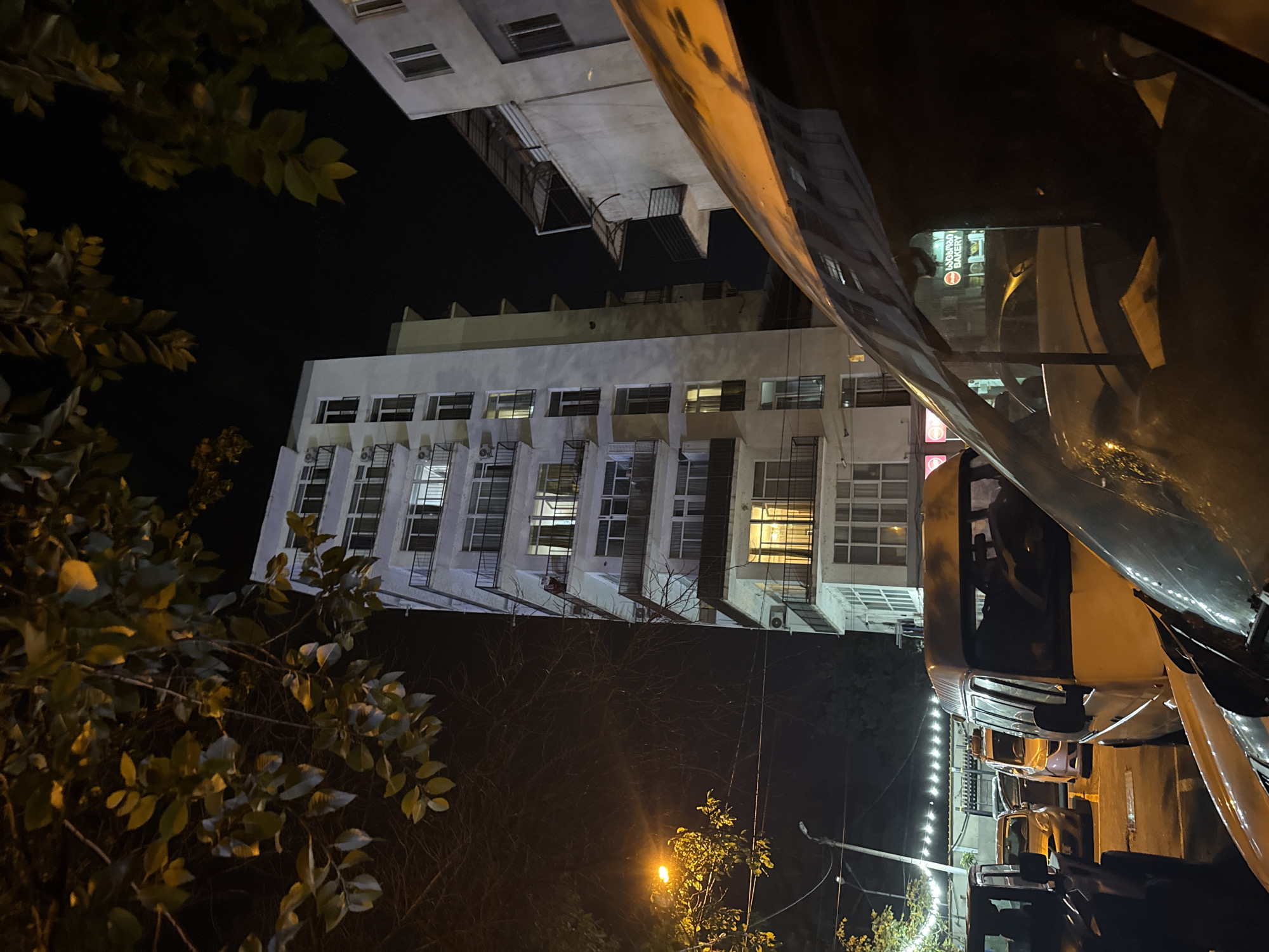 In Tbilisi, Georgia, at night, a tall, multi-story white building with numerous windows and balconies stands prominently. The building is illuminated by artificial lights, with some windows glowing warmly from within. In the foreground, the hood and windshield of a vehicle are visible, reflecting the surrounding environment. The scene includes several parked vehicles, including vans and cars, on what appears to be a street or parking area. Trees with visible leaves frame the top of the image, and a street lamp casts a yellowish light. The overall atmosphere is urban and nighttime, with a mix of residential and commercial elements suggested by the building and the presence of businesses, one of which displays a sign with Georgian script and the word "BAKERY."
