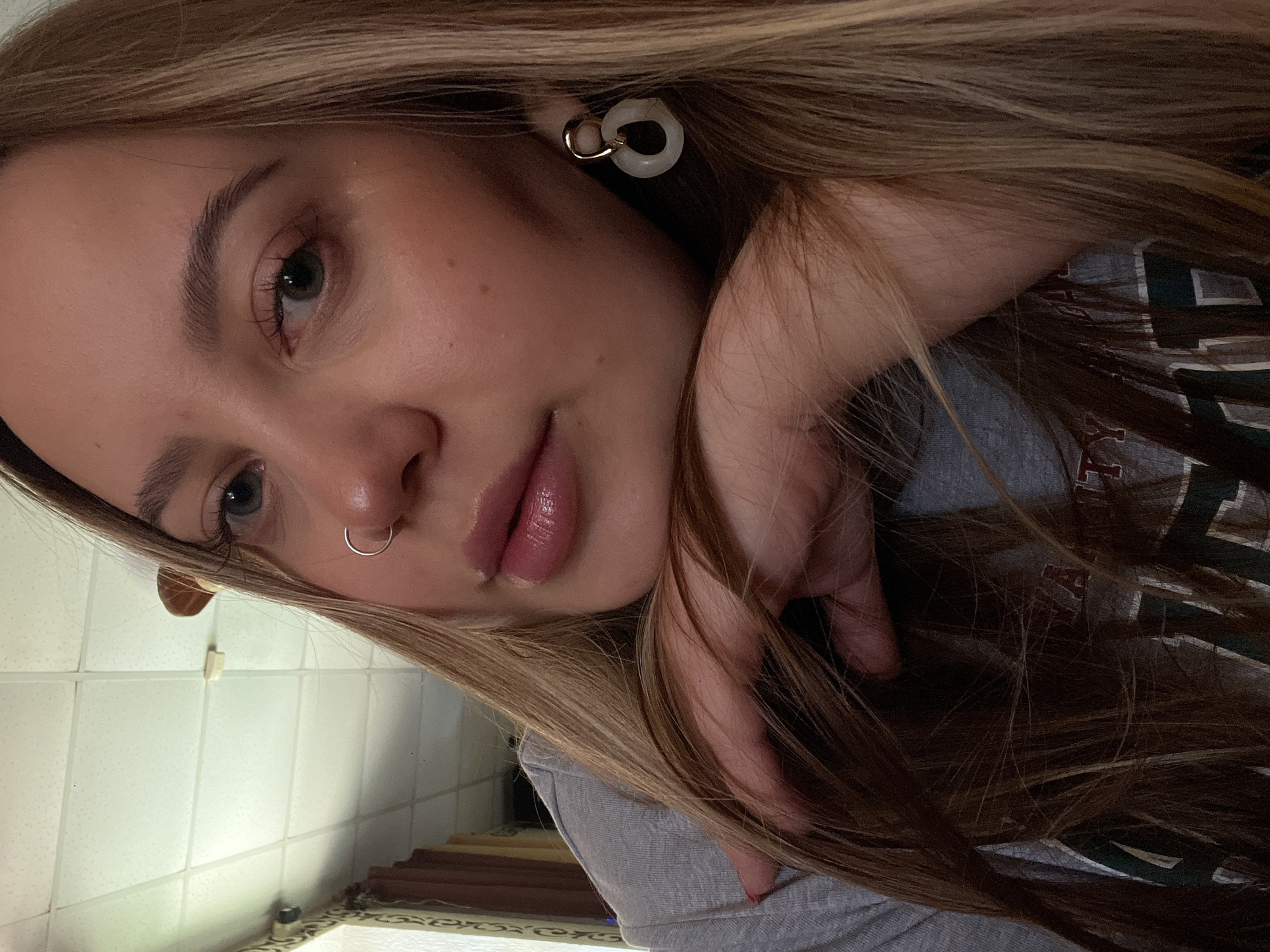 A young adult female is shown in an indoor close-up portrait, resting her chin on her right hand, which is mostly concealed by her long, straight brown hair with blonde highlights. She has blue-green eyes, defined eyebrows, and a silver hoop nose ring in her left nostril. Her lips are full and adorned with glossy, light pink lipstick. She wears a grey t-shirt displaying a partial dark green and white graphic design. On her right ear, she has an earring featuring a gold-toned hook attached to an irregularly shaped white circular pendant. The background primarily consists of reflections, likely from a mirror, revealing a white grid-paneled ceiling with a visible light fixture or vent, and a textured wall section. This scene is situated in an interior location within Novopokrovskaya, Russia.