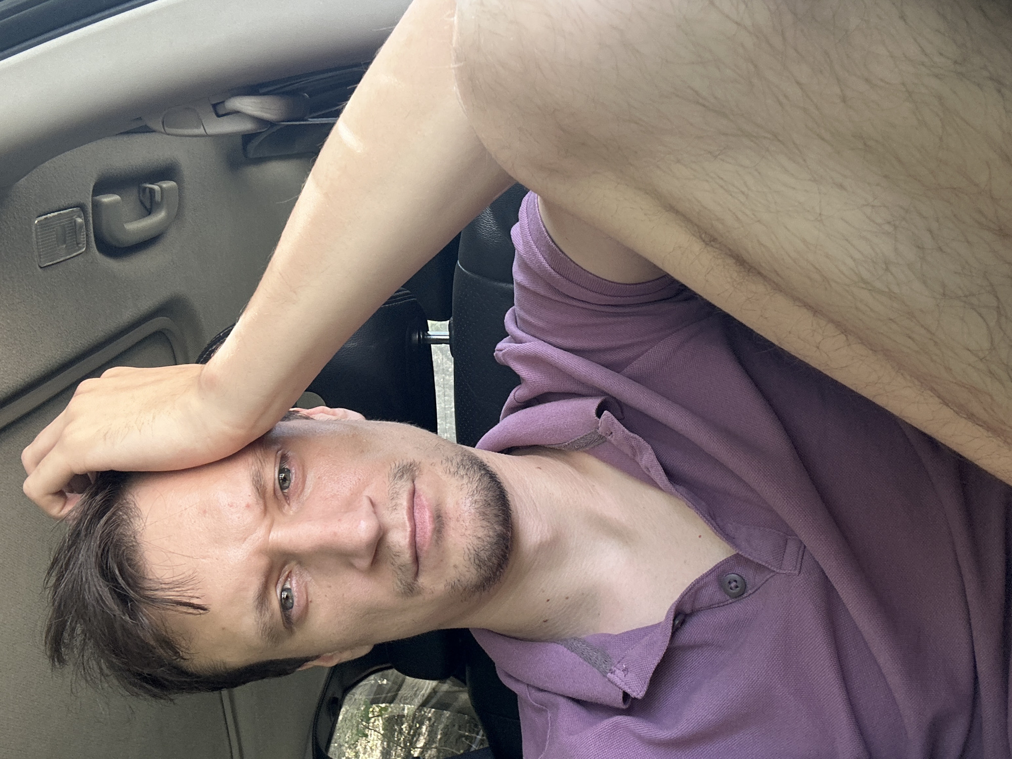 A young adult male with light brown hair and a goatee is sitting in the passenger seat of a vehicle. He is wearing a purple polo shirt and has his right hand on his forehead, with his arm bent. The background shows the interior of the car, including the ceiling, a handle, and part of a seat. The male appears to be looking at the camera with a serious expression. The image does not provide any information or context about the location in the city of Makiv, Ukraine.