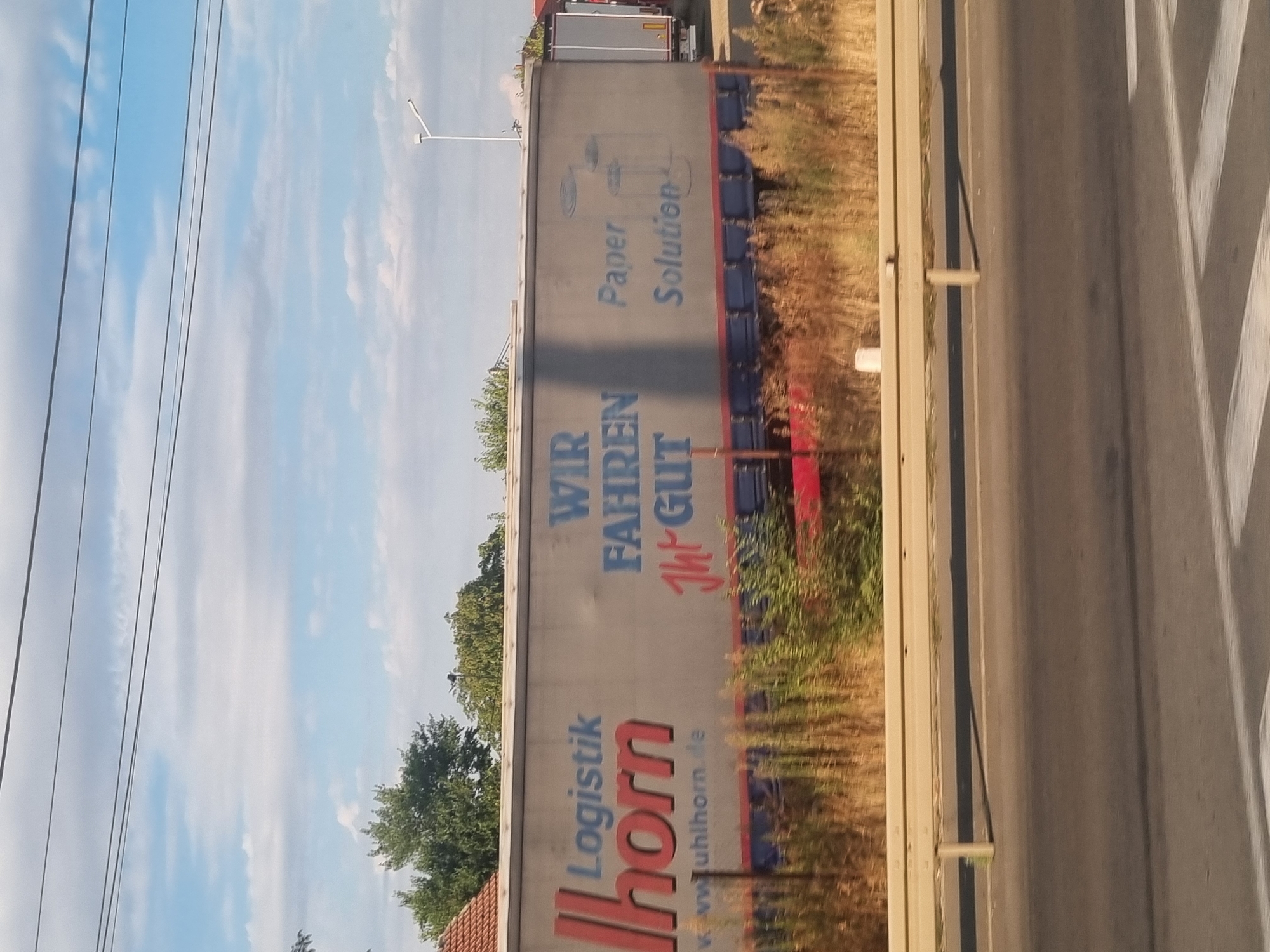 The image shows a side view of a large trailer truck parked along a road. The truck is primarily gray with blue and red lettering. On the side of the trailer, there is a prominent logo for "Ilhorn Logistik" in large red and blue text, with a website address "w. w.uhlhorn.de" below it. Further to the right, in blue text, is the phrase "WIR FAHREN GUT" and beneath that "Paper Solution". There is also a sketch of what appears to be a paper shredder or recycling bin. The truck is parked on a paved surface, possibly a road or parking lot, with dry, tall grass and vegetation in the foreground. A yellow guardrail runs along the side of the road. In the background, there are trees and a clear blue sky with scattered clouds. The overall scene suggests an industrial or logistical area.