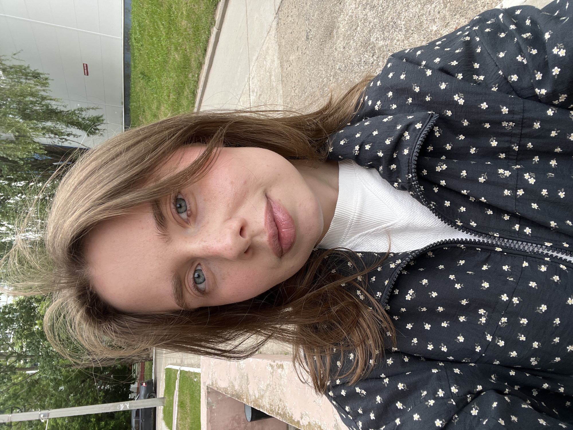 This image is an outdoor selfie of a young woman, rotated 90 degrees counter-clockwise. The woman has light brown, shoulder-length hair and blue-grey eyes, looking directly at the camera with a neutral to slightly pleasant expression. Her fair complexion shows some freckles on her cheeks, and her lips are slightly parted with a natural pink tone.
She is wearing a dark-colored (likely black or dark blue) jacket or top, patterned with small white and light yellow flowers. A white, ribbed or textured garment is visible underneath.
The background suggests an urban or suburban environment, consistent with Chaikovsky, Russia. To her left (top in the rotated image), there's a light-colored building facade featuring a red rectangular sign or object, the text of which is unreadable. Green grass, trees, and a paved area are also visible. To her right (bottom in the rotated image), the scene includes more green grass, a paved surface, and a concrete ledge or barrier. Further back, additional trees and a dark-colored car are partially visible.
The scene is set during the daytime, with soft, diffused lighting that suggests an overcast sky, as no harsh shadows are present. The activity is a casual self-portrait.