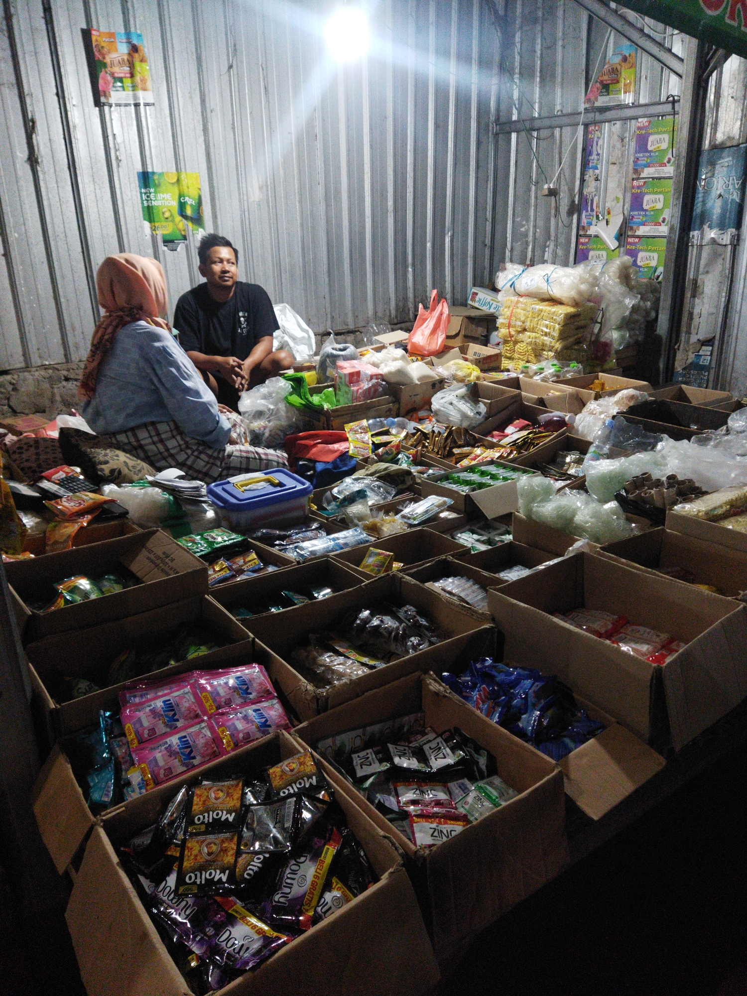 This image captures a scene inside a modest stall in Semarang, Indonesia, likely a street-side market or a small shop. Two individuals are present: a woman wearing a light blue hijab and a plaid skirt, seated on the ground, and a man in a black t-shirt, also seated. They appear to be interacting or perhaps waiting for customers in the dimly lit environment, illuminated by a bright light source from above.
The stall is filled with an assortment of goods displayed in cardboard boxes. The boxes are arranged on the floor and seem to contain various packaged products, including what appear to be snacks, cleaning supplies, or small household items. Some of the packaging is brightly colored and features text in Indonesian. Visible items include packets of what look like coffee or creamer, shampoos, and possibly snacks like crackers or cookies. In the background, the walls are corrugated metal, a common material for temporary structures in such markets. Posters are affixed to the walls, advertising products and bearing local language text. The overall impression is of a small, functional retail space with a variety of goods for sale.