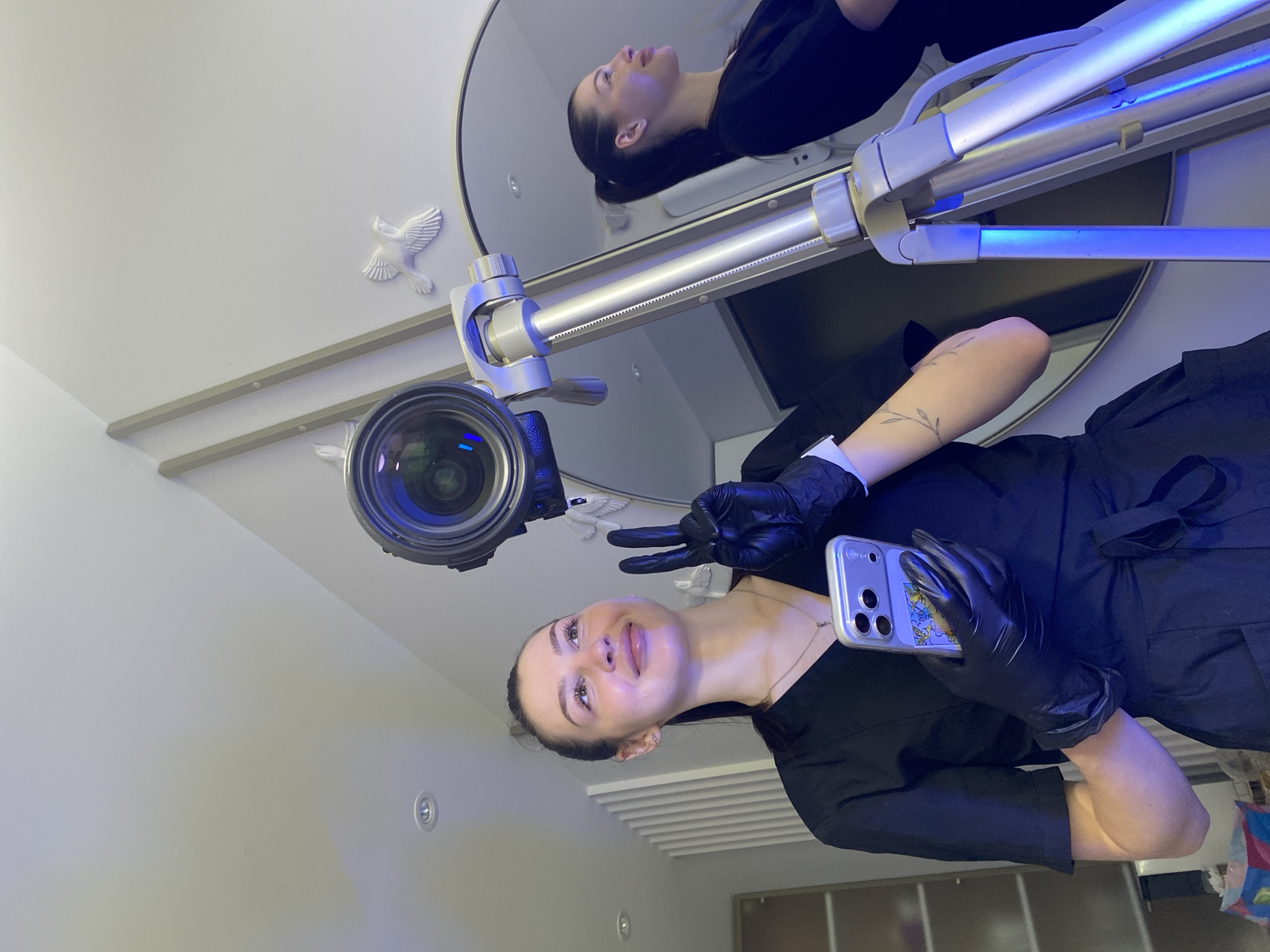 A woman wearing a black shirt and black gloves makes a "peace" sign with her hand while holding a cell phone. The photo is taken from a low angle, looking up towards her. A large camera lens is mounted on a metal arm next to her head. Behind her, a round mirror reflects another woman lying down. The room appears to be brightly lit, with white walls and ceiling. There are decorative bird figures on the ceiling near the mirror. The photo was taken in Votkinsk, Russia.