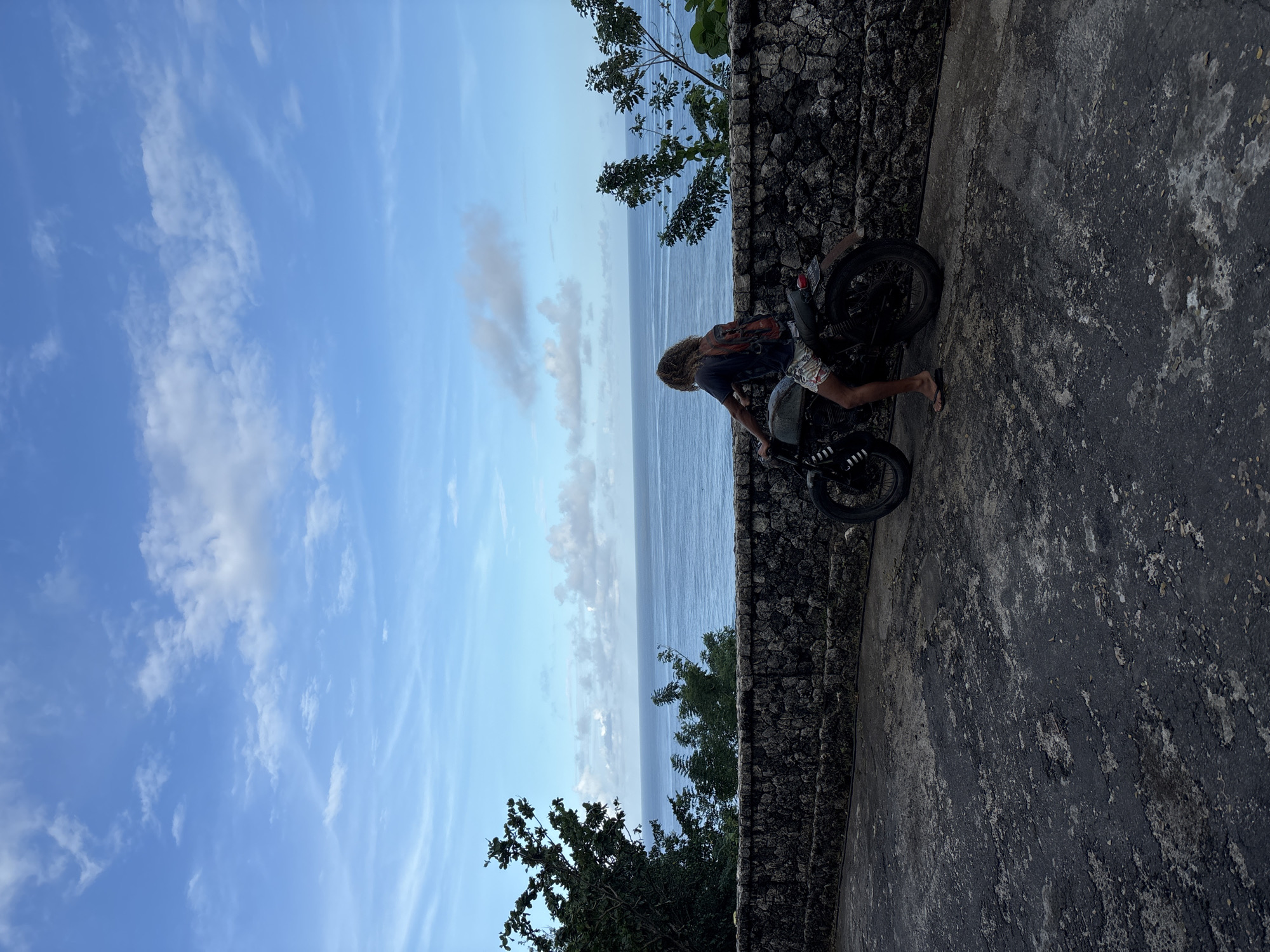 A man with curly hair wearing a dark shirt, light-colored shorts, and sandals is standing on a concrete ledge next to a stone wall. He is beside a black motorcycle. Beyond the wall is the ocean. The sky is blue with some clouds. The location is Kuta Selatan, Indonesia.