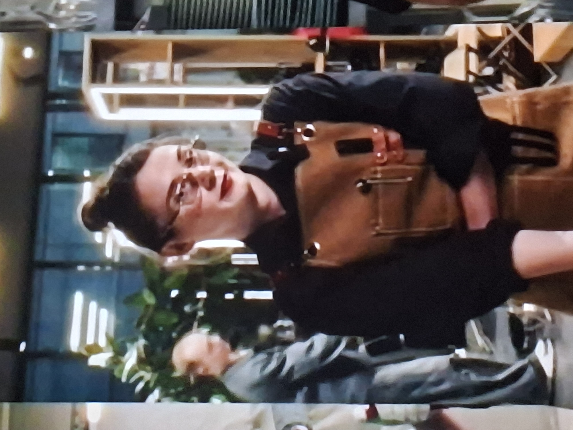 The image shows two individuals in what appears to be a workshop or studio setting. The primary subject is a person, possibly female, wearing glasses and a black shirt, with their hair styled in a bun. They are also wearing a brown apron with straps that have buckles. This person is leaning forward, with their head tilted, and their mouth is open as if speaking or reacting. Their expression seems animated.
Behind this individual, another person is partially visible, lying down or reclined. This second person is wearing a gray jacket or top.
The background suggests an indoor environment with shelves filled with various objects, including what look like tools or materials. There are also hints of greenery from plants in the lower left portion of the image. The lighting suggests it might be daytime, with artificial lights also present, indicated by bright rectangular reflections in the background.
The overall scene conveys a sense of activity within a creative or making space. There is no visible text in the image. The context provided indicates the location is Ivanovo, Russia.