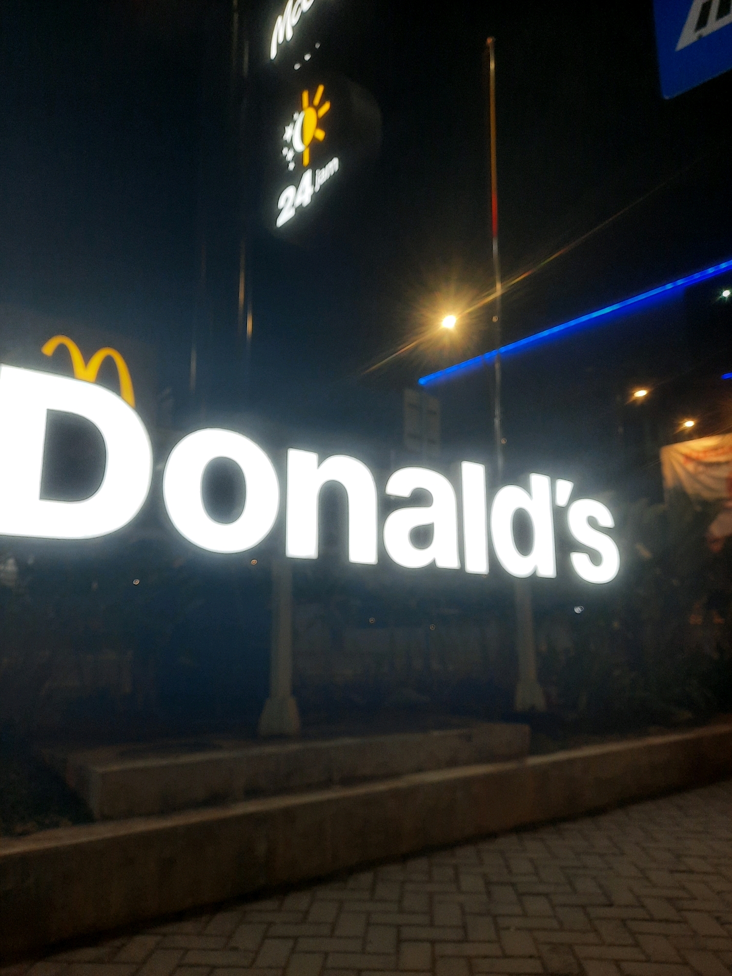 A large, illuminated "McDonald's" sign is prominently displayed at night in North Jakarta, Indonesia. The sign features the iconic golden arches logo above the white, illuminated text "McDonald's". To the left of the main sign, a smaller illuminated sign for "McDonald's" indicates "24 jam" (24 hours) and displays a sun and moon graphic. The signage is erected on a concrete structure, with patterned brick paving visible in the foreground. The background is dark, with some blurry lights and structures, suggesting an urban environment. There are no people visible in the image.