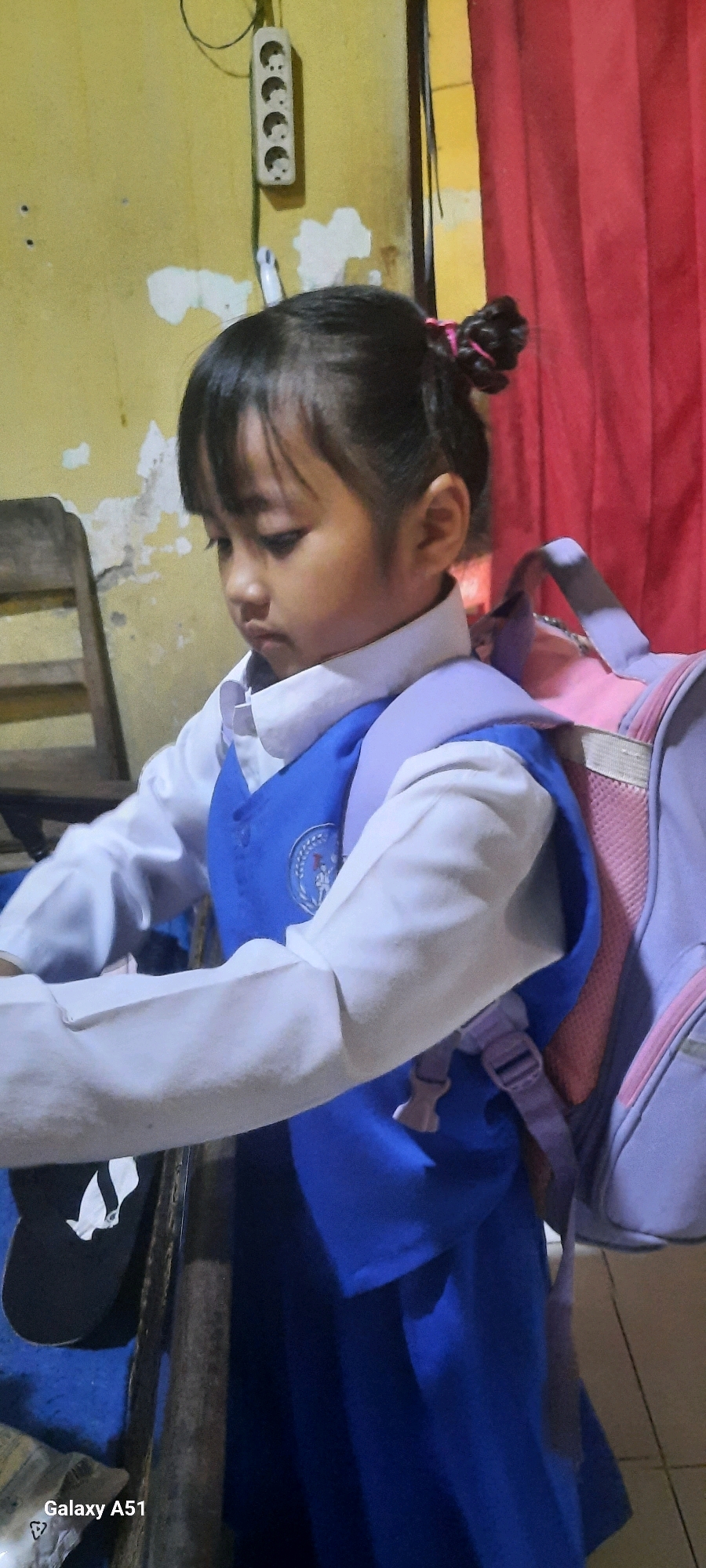 A young girl with dark hair pulled into a bun is wearing a white shirt and a blue vest. She has a pink and lavender backpack on her shoulders. The background is a yellow wall with some white plaster peeling off and a red curtain on the right. A white electrical outlet with four sockets is mounted on the wall. A wooden chair is visible on the left. The scene appears to be indoors, possibly in a home or school in Ploso, Indonesia, given the watermark at the bottom left.