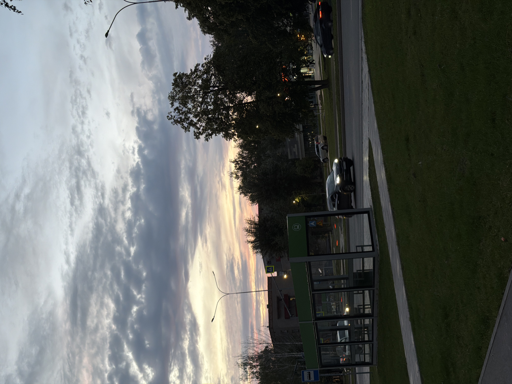 This is a photograph taken in Nizhnekamsk, Russia, during dusk. The sky is filled with dramatic clouds, displaying hues of gray, white, and soft pinks, indicating the setting sun. The scene is urban, with a road running alongside a grassy area and a sidewalk.
Prominent objects include a modern, green bus stop shelter with glass panels, a street lamp with a distinctive curved design, and several cars driving or parked along the road. Some buildings are visible in the background, along with trees that are partially silhouetted against the colorful sky.
The activity appears to be the normal flow of a city street at the end of the day. There are no visible people in the frame, but the presence of a bus stop and cars suggests ongoing urban activity. The lighting suggests it is either early morning or late evening, with the soft, diffused light creating a peaceful atmosphere. The weather appears clear, with the sky dominated by the cloud formations.
There are some signs visible, including a blue bus sign on the bus stop and yellow warning signs on poles along the road, indicating pedestrian crossings. The overall impression is a quiet, urban scene bathed in the soft light of dusk.