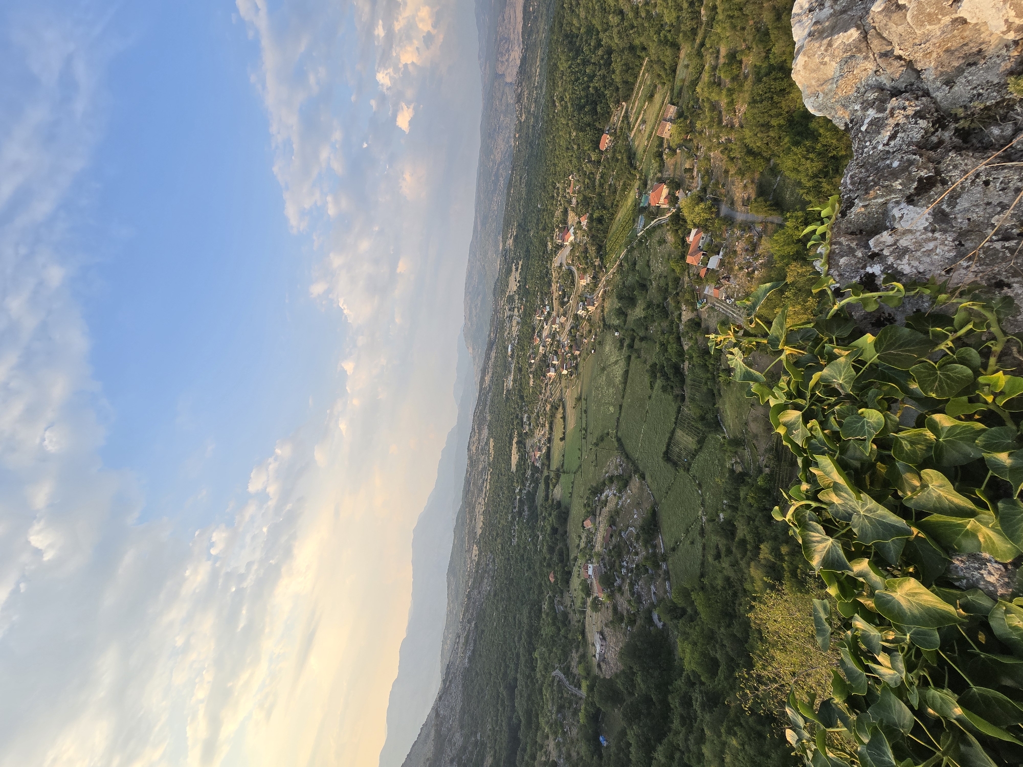 The image depicts a panoramic view of the landscape surrounding Medun, Montenegro, as seen from a high vantage point. In the foreground, a rocky outcrop is partially covered by lush green ivy, with sunlight highlighting the leaves. The middle ground features a valley dotted with scattered houses with red-tiled roofs, interspersed with green fields and dense trees. A winding road is visible, connecting the houses. The background is dominated by rolling hills and distant mountains, fading into a hazy horizon. The sky above is a mix of blue with scattered white and gray clouds, with the warm glow of late afternoon or early morning sunlight illuminating the scene from the left. The overall impression is one of rural tranquility and natural beauty.