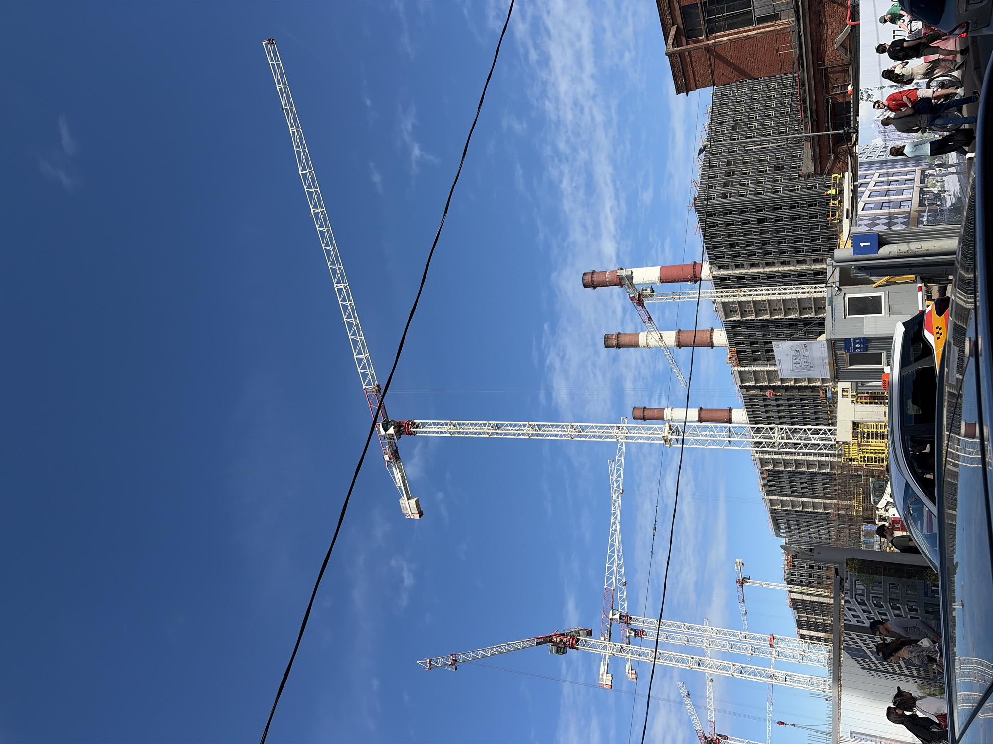 The image depicts a construction site in St. Petersburg, Russia, under a clear blue sky with scattered clouds. Several tall, white construction cranes dominate the skyline, with their long booms extending across the frame. In the background, a large, multi-story building is under construction, its skeletal structure visible with numerous windows. Adjacent to the new construction are two tall red and white smokestacks, remnants of an industrial past. The foreground shows a street scene with cars and pedestrians. People are walking and standing near a construction fence adorned with architectural renderings of the future buildings. A silver car is parked in the lower portion of the frame, reflecting the sky. The overall scene captures a transition between old industrial infrastructure and modern urban development in St. Petersburg.