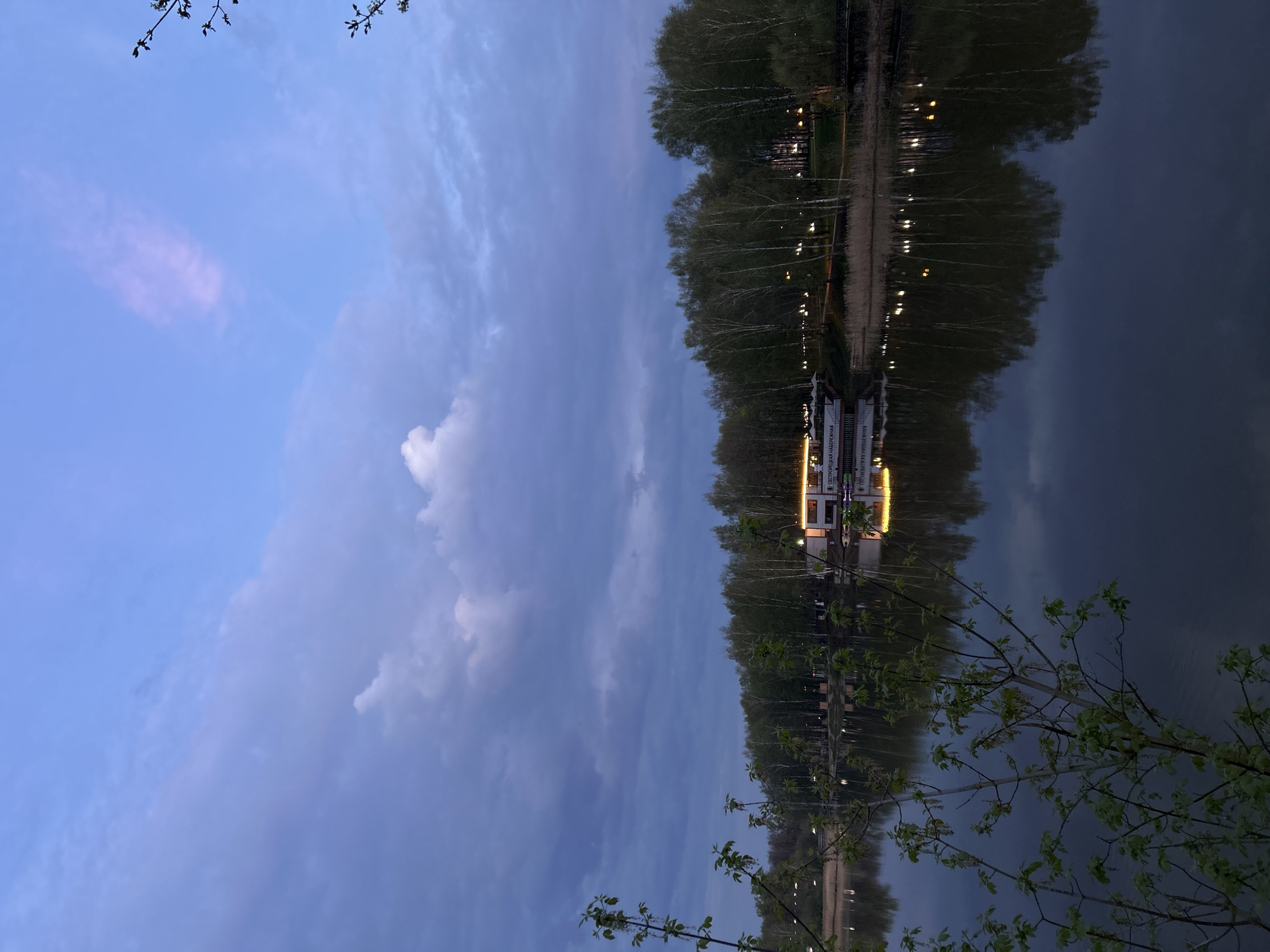 The image depicts a tranquil scene at night, showing a body of water reflecting the surrounding environment. Along the water's edge, trees line the shore, their reflections mirrored in the calm surface of the water. Lights illuminate the area, creating a warm glow that is also reflected in the water. In the distance, a structure is visible with more lights, suggesting a building or establishment along the waterfront. The sky above is a gradient of blues and grays, with clouds scattered across the horizon. Branches with budding leaves frame the bottom right corner of the image, adding a touch of nature to the composition. The location is Klin, Russia, based on the text on the boat.