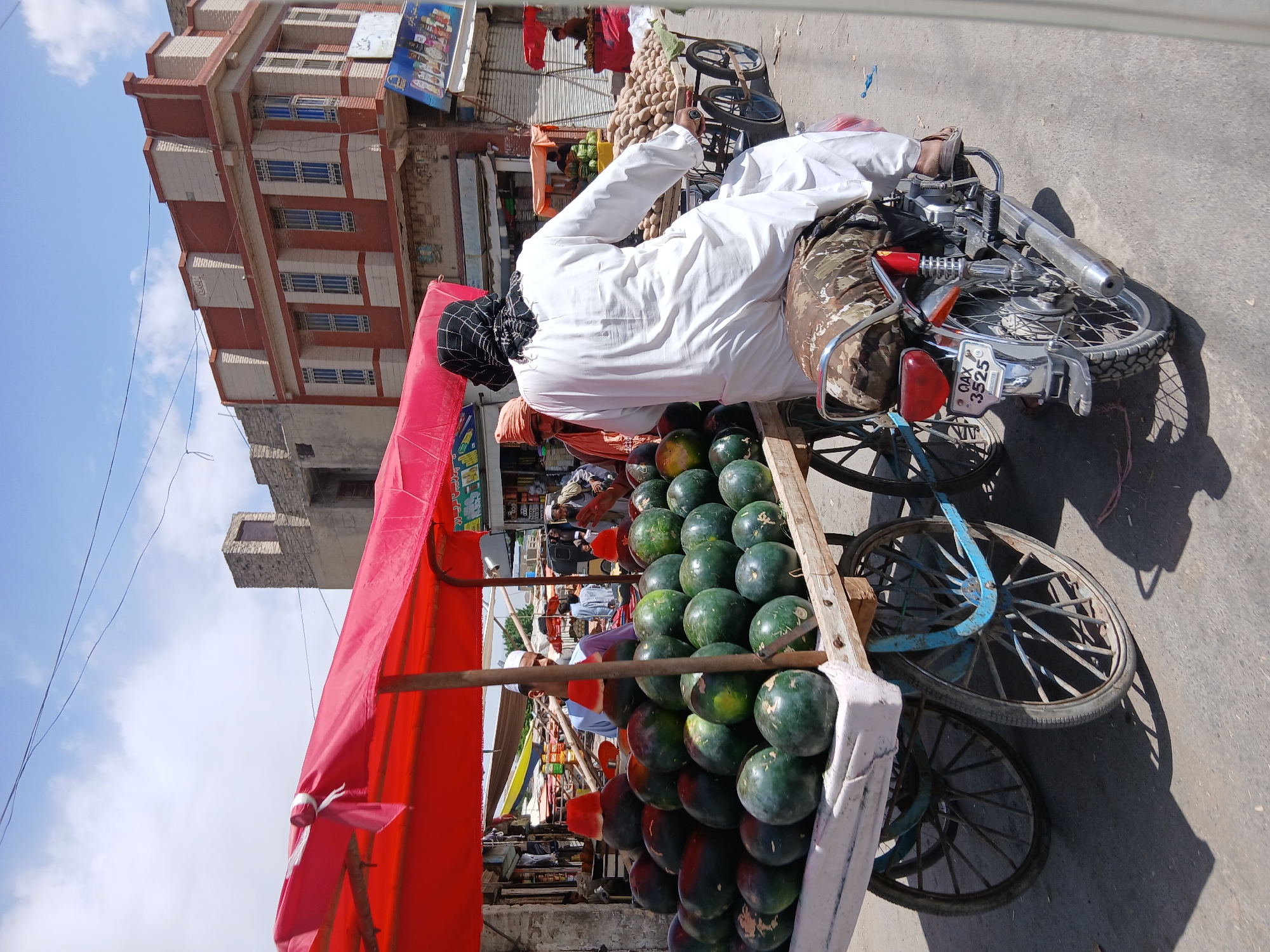 A man wearing a white garment and a black and white head covering is sitting on a motorcycle attached to a cart loaded with watermelons. The cart has a red canopy. The motorcycle's license plate reads "QAx 3525." Behind the cart, a row of shops lines the street. In the background, there's a multi-story building with a red and white facade. The photo was taken outdoors under a bright sky with some visible clouds, suggesting daylight. The setting appears to be a street market, likely in Quetta City Tehsil, Pakistan.