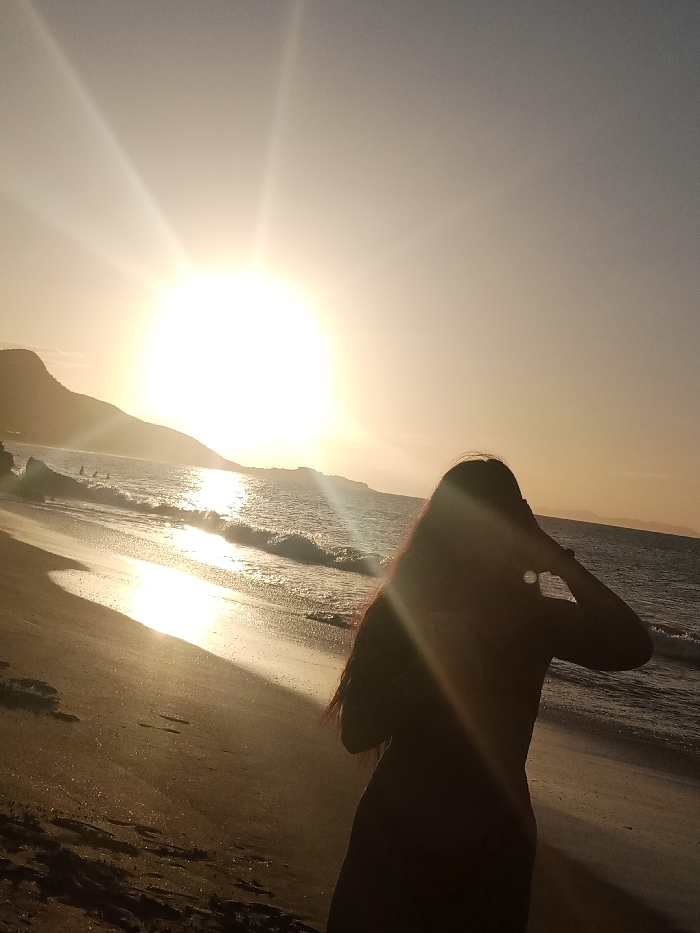 A beach scene features a silhouetted figure of a woman with long hair standing on the sand, positioned with her back to the camera. The sun is setting in the background, creating a bright, golden glow across the sky and reflecting off the ocean waves. A silhouette of a mountain or landmass is visible on the left horizon, completing the scenic backdrop. The overall tone is warm and serene, emphasizing the beauty of a sunset at the beach.