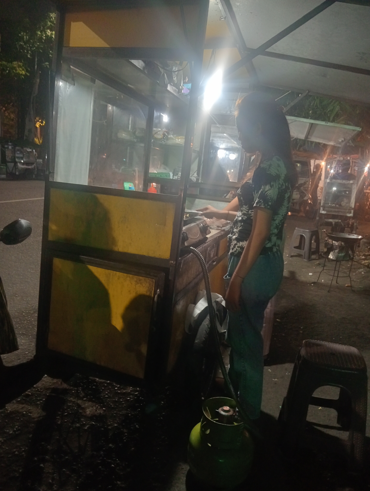 A woman in a floral print shirt and teal pants stands next to a street food cart in Surabaya, Indonesia. The cart is yellow and appears to be made of metal and glass. A gas cylinder is visible on the ground in front of the cart. In the background, there are other food stalls, stools, and parked motorcycles, indicating a busy street market or roadside food area. The lighting suggests it is nighttime.