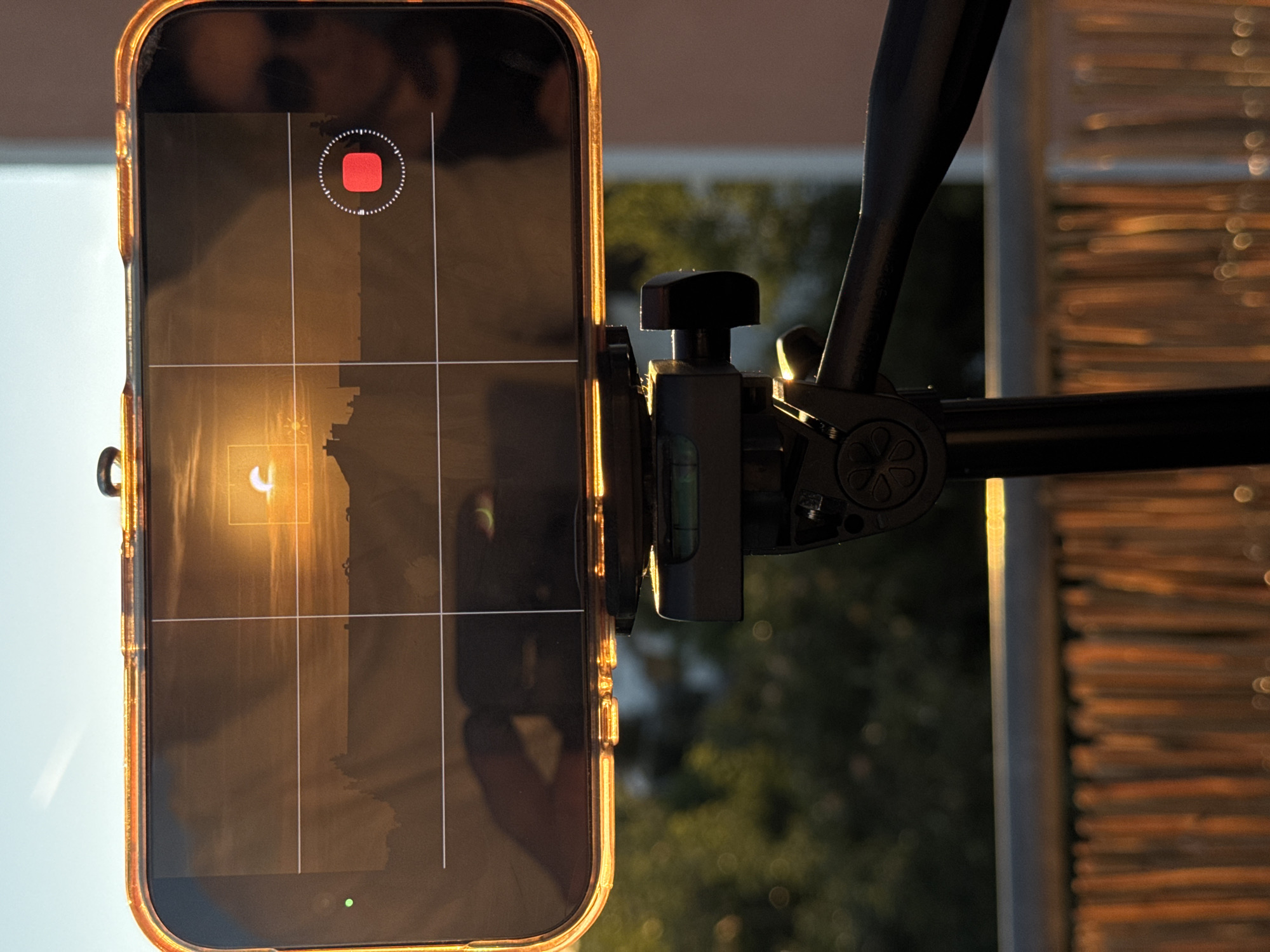 A smartphone is mounted on a tripod, displaying a camera interface. The screen shows a sunrise or sunset over a cityscape, with a partial solar eclipse visible. The phone's case is transparent with an orange tint. A red record button is illuminated on the screen. The tripod is positioned on a balcony or rooftop, with a blurred background of greenery and a building. The foreground shows a railing and a wooden fence. The image is taken outdoors in Wettstetten, Germany.