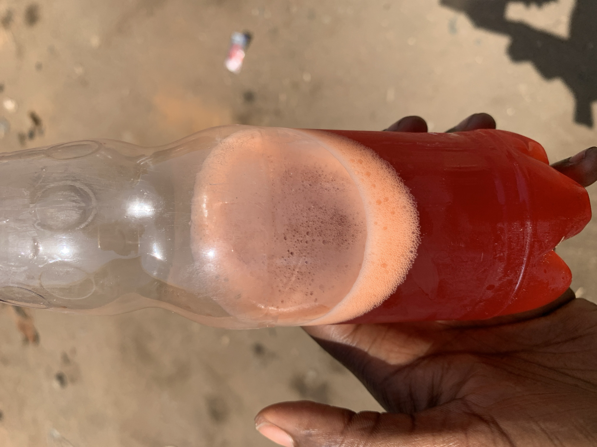 A hand holds a horizontally oriented, partially filled plastic bottle. The bottle contains two distinct liquids: a clear liquid occupies roughly half the bottle, while the remaining half is filled with a frothy, reddish-orange liquid. The hand is dark-skinned. The background shows a rough, light-brown surface and a blurred shadow. A discarded bottle is visible in the top left of the frame. The location is Rafin Par, Nigeria.