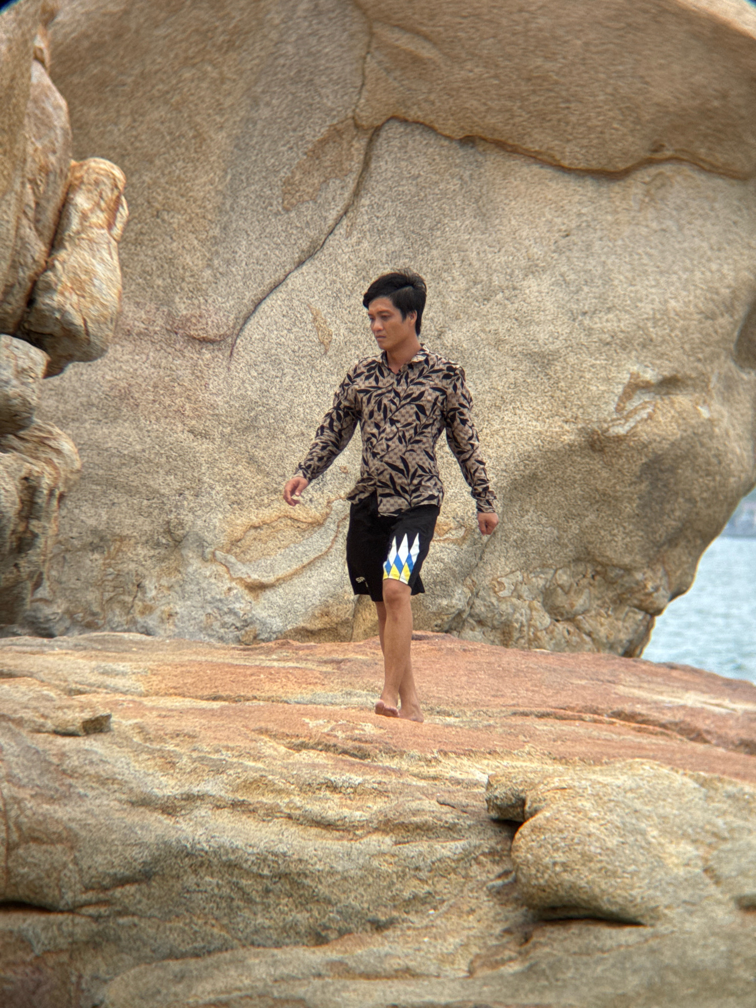 An East Asian male, appearing to be in his early twenties, is walking on a rocky shore, likely in Nha Trang, Vietnam. He is wearing a long-sleeved patterned shirt with a dark floral print on a light brown background, and black shorts with a geometric pattern of blue, yellow, and white diamonds. The man is looking down and to his right. The environment consists of large, weathered rocks in shades of beige and tan, with the ocean visible in the background. The lighting suggests it is daytime, possibly late afternoon given the warm tones of the rocks. The texture of the rocks indicates they are exposed to the elements and have been shaped by erosion.