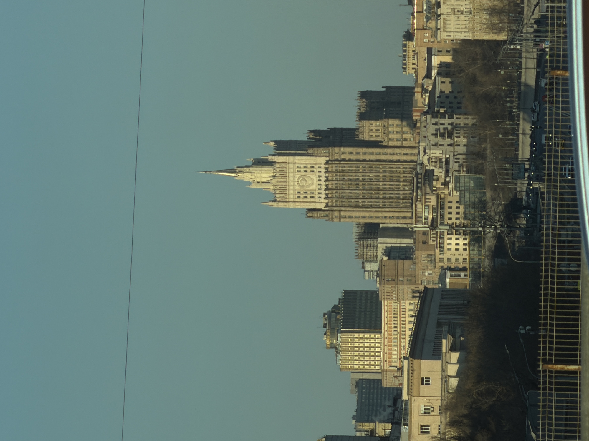 The image shows a view of the Ministry of Foreign Affairs building, one of the Seven Sisters Stalinist skyscrapers, located in Moscow, Russia. The building's spire and clock face are visible against a blue sky. Other buildings and trees are also visible in the cityscape. A window and frame partially obscure the right side of the image.