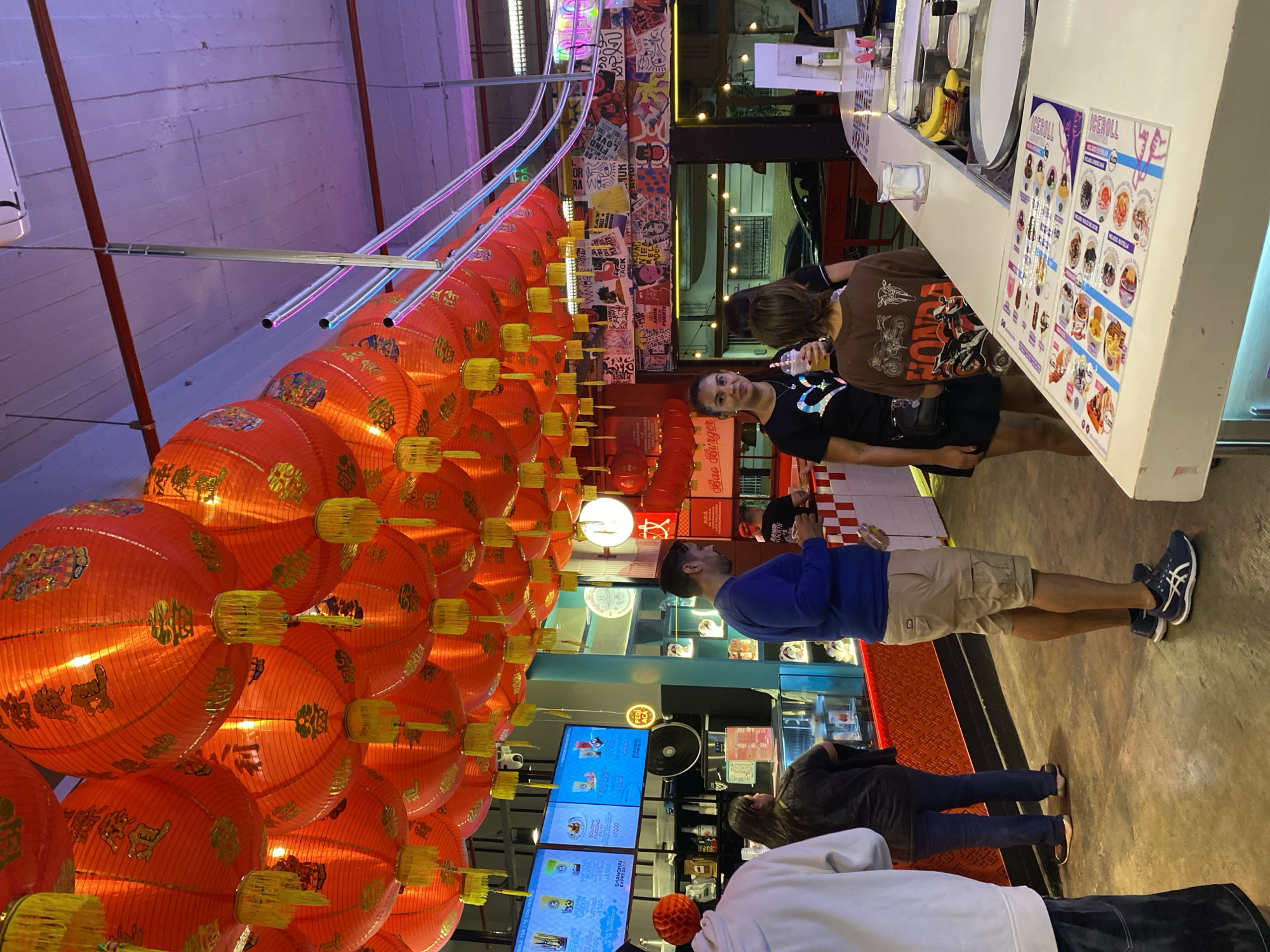 Here's a forensic description of the image:
**Scene:** Indoor food establishment. The view is angled upwards, showcasing a ceiling crowded with strings of Chinese red lanterns. Fluorescent lighting fixtures run parallel to the lantern strings. Walls are decorated with a mix of posters and graphics.
**People:** Several individuals are present:
* One woman is looking towards the camera.
* A man in a blue shirt and khaki shorts is standing near a counter, likely a customer.
* Other customers are present.
**Objects:**
* Red Chinese lanterns hang in rows.
* A counter serves as a food preparation and ordering area.
* Menus are visible.
* Refrigerated display cases with food and drinks are behind the counter.
* Digital screens show food and beverage options.
**Actions/Interactions:**
* Customers are likely ordering food or beverages.
* The person near the counter is engaged with the food ordering.
**Environment:**
* The location is an enclosed space.
* The atmosphere is dimly lit with colorful lights.
* Based on signage, the store name is related to "Bao Burger".
**Location:**
* Signs indicate the location is in Buenos Aires, Argentina, based on store names.