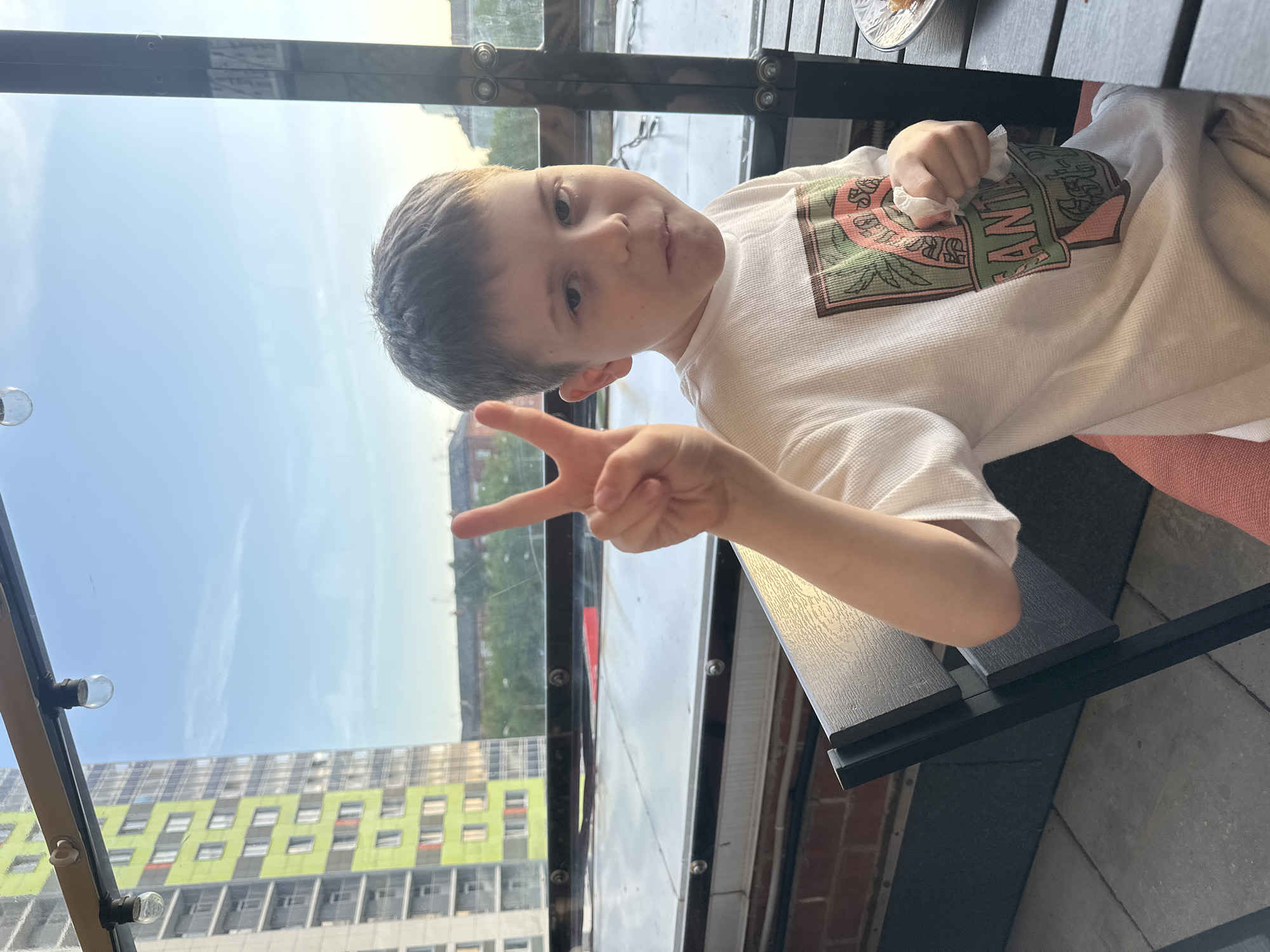 The image shows a child making a peace sign gesture. The child is wearing a white shirt with a graphic design. They appear to be indoors, possibly in a restaurant or enclosed space with a view. Outside, there is a view of an apartment building with a modern design, featuring green and white sections. Based on the location, specifically the presence of similar buildings, this image was likely taken in the city of Perm, Russia.