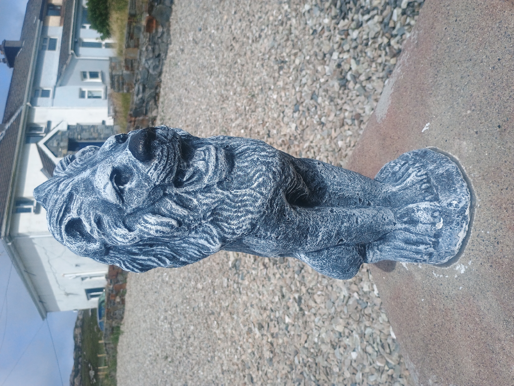 An outdoor, eye-level, medium shot captures a stone lion statue positioned on a light brown, textured surface. The statue, predominantly gray with white accents, faces right, with its head turned forward. It is seated with its front paws touching the ground. The statue is the primary subject, filling the lower half of the frame.
Behind the statue, the ground transitions to a light gray gravel surface. Further in the background, a low stone wall runs horizontally across the image, separating the gravel from a grassy area. Beyond the wall, a white house with a gray roof and multiple windows is visible. To the far left of the background, a green, hilly landscape stretches under a bright, clear blue sky with scattered white clouds. A silver car is parked on a gravel driveway in front of the house. The lighting suggests it is daytime, with sunlight casting faint shadows.