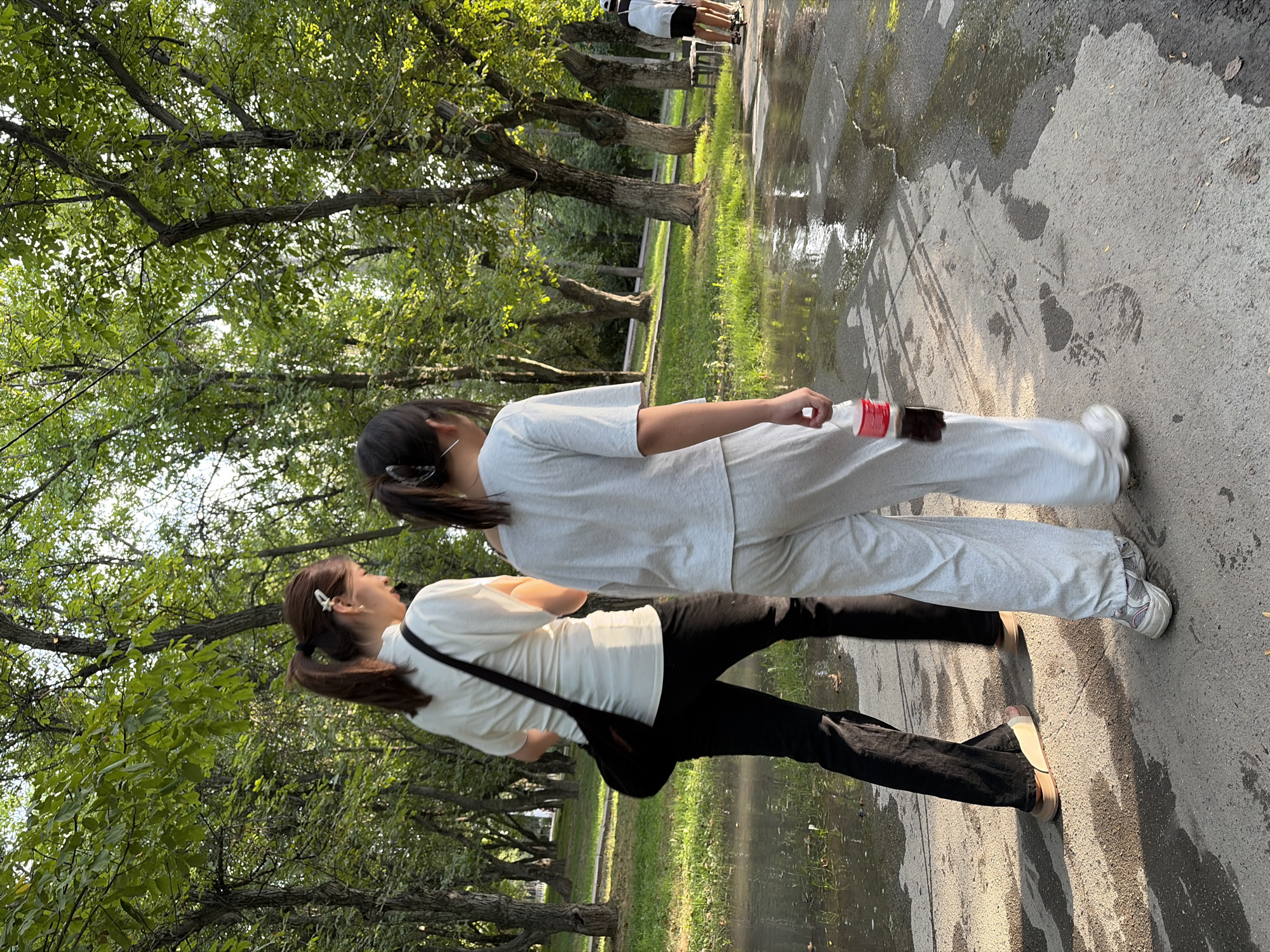 In an outdoor park setting in an unknown city and unknown location, two individuals are walking on a wet paved pathway. The person on the left, facing away from the camera, is wearing a white short-sleeved shirt, black pants, and tan sandals. They have a dark-colored bag slung over their left shoulder. The person on the right, also facing away, is wearing a light gray t-shirt and light gray sweatpants. They are holding a red and clear plastic bottle with a dark liquid in their right hand. The ground is wet with puddles and has some footprints visible. Lush green trees and grass surround the pathway, indicating a natural environment within the city. In the background, further down the path, another individual wearing white and black is visible, appearing to be on a scooter. The lighting suggests it is daytime, possibly late afternoon, with sunlight filtering through the trees.
