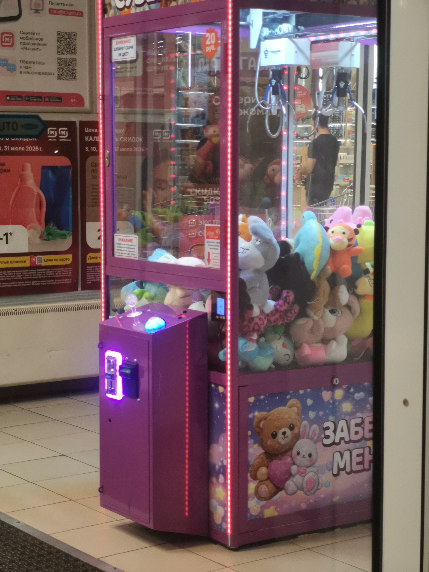 The image shows a claw machine filled with plush toys, situated inside a commercial establishment, likely a shopping mall or supermarket, as indicated by the tiled floor and signage. The machine is primarily purple and illuminated with pink LED lights. A sign on the machine reads "20 руб." (20 rubles), indicating the price per play. There are various plush toys inside, including a light blue dinosaur, a light blue elephant, a pink bunny, and a light orange tiger. A portion of a person wearing a dark t-shirt is visible inside the machine, appearing to operate it. The overall environment suggests a retail space. The image does not contain specific details to confirm its location in Cherkessk, Russia.