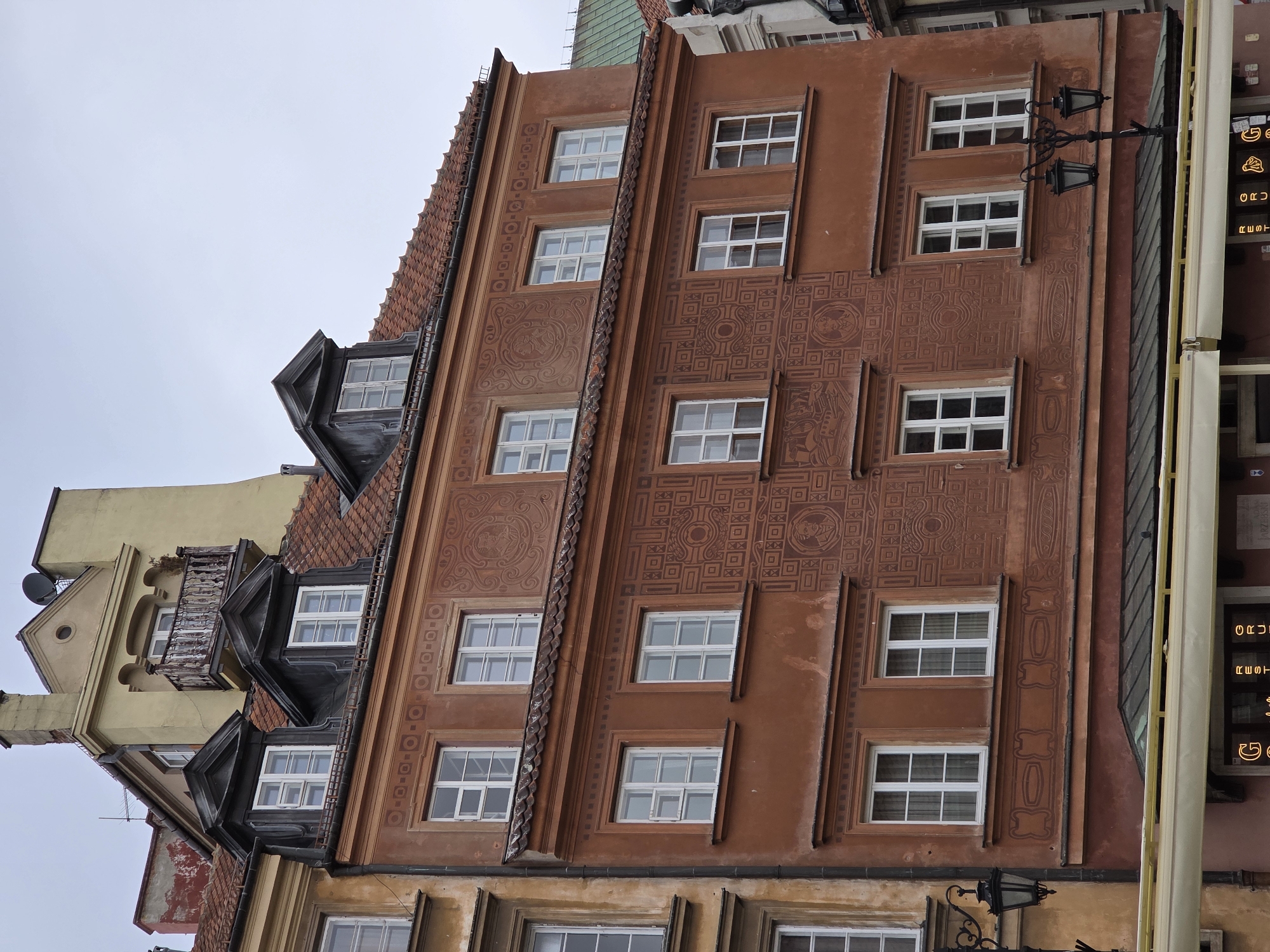 The image shows a view of buildings in the city of Warsaw, Poland. The buildings are historic and ornate, with multiple windows and detailed facades. The architecture is reminiscent of the Old Town area of Warsaw. The buildings appear to be of various colors, including brown, yellow, and terracotta. The sky is overcast, and the image is shot from a low angle. There are no people visible.