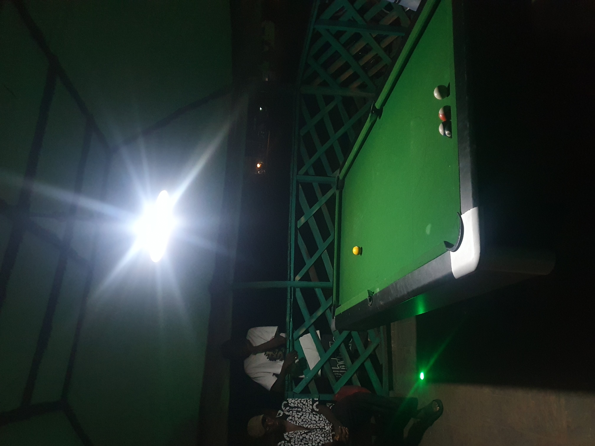 In Lokoja, Nigeria, a dimly lit outdoor establishment features a pool table as its centerpiece. A bright light source overhead creates a lens flare, illuminating the green felt of the pool table. Three pool balls are visible on the table: a solid red ball with a number, a striped ball with a number, and a solid white ball. To the left, two men are present. One man, wearing a cap and a patterned shirt, is seated, while another man in a white t-shirt stands leaning on a green railing. A small green light is visible on the ground near the seated man's feet. The environment appears to be an open-air structure with a green, lattice-style railing.