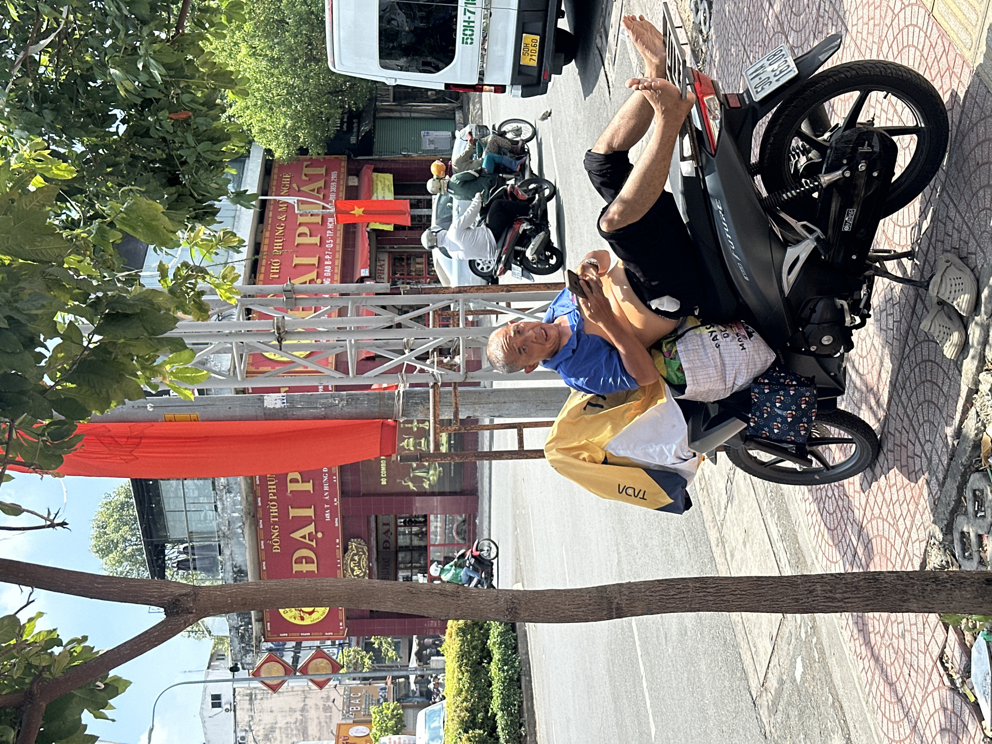 A man is reclining on a parked motorcycle on a sidewalk in Thủ Đức, Vietnam. The motorcycle has license plate number "50-AA 166.08". He is holding a smartphone and looking toward the camera. He wears a blue shirt and dark shorts. Bags are hanging on the motorcycle. A van with license plate "50H-710.60" is parked behind the motorcycle. Other motorbikes are visible on the street. Buildings with red signs are in the background, including one that reads "ĐẠI P" and another "ÁI PHÁT" with the phone number "(08) 3859.2985". A Vietnamese flag is displayed. Street signs and trees are also present.