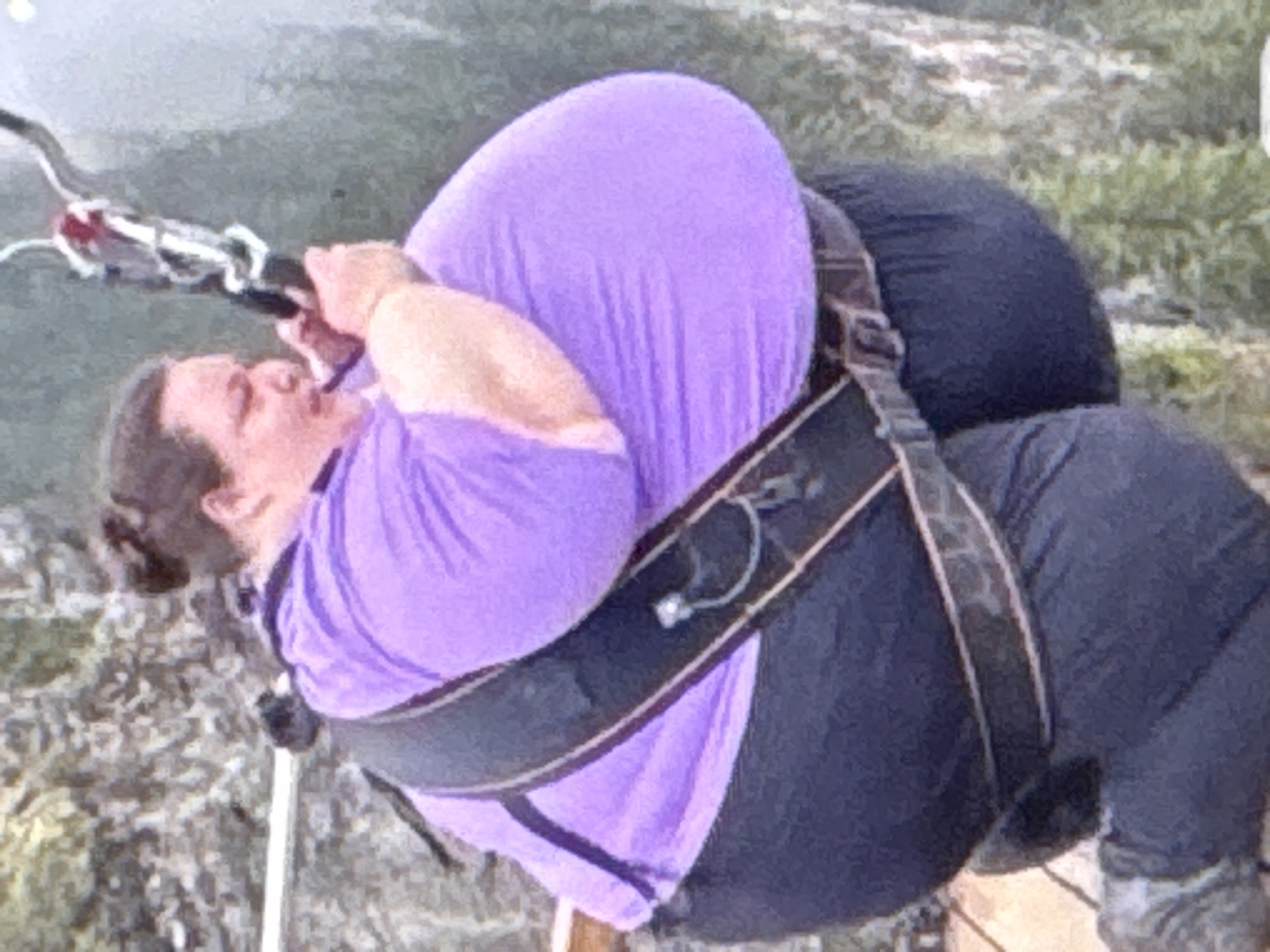 The image shows a person dressed in a purple t-shirt and dark pants, secured by a harness. The person's body is largely obscured by the clothing and harness, but it appears they are in a reclined or suspended position. The background is blurred, suggesting a natural outdoor environment with foliage. The lighting indicates it may be daytime, with no strong shadows. There is no visible text. The image is a close-up and tilted, focusing on the person and their equipment. The scene appears to be outdoors, possibly in a mountainous or hilly area given the background texture. The person's face is visible and appears to be strained or pained. The context of Kuta Selatan, Indonesia, may suggest a tropical environment, though this is not directly evident from the image itself. The activity could be related to adventure sports or a rescue situation, given the harness and suspension.