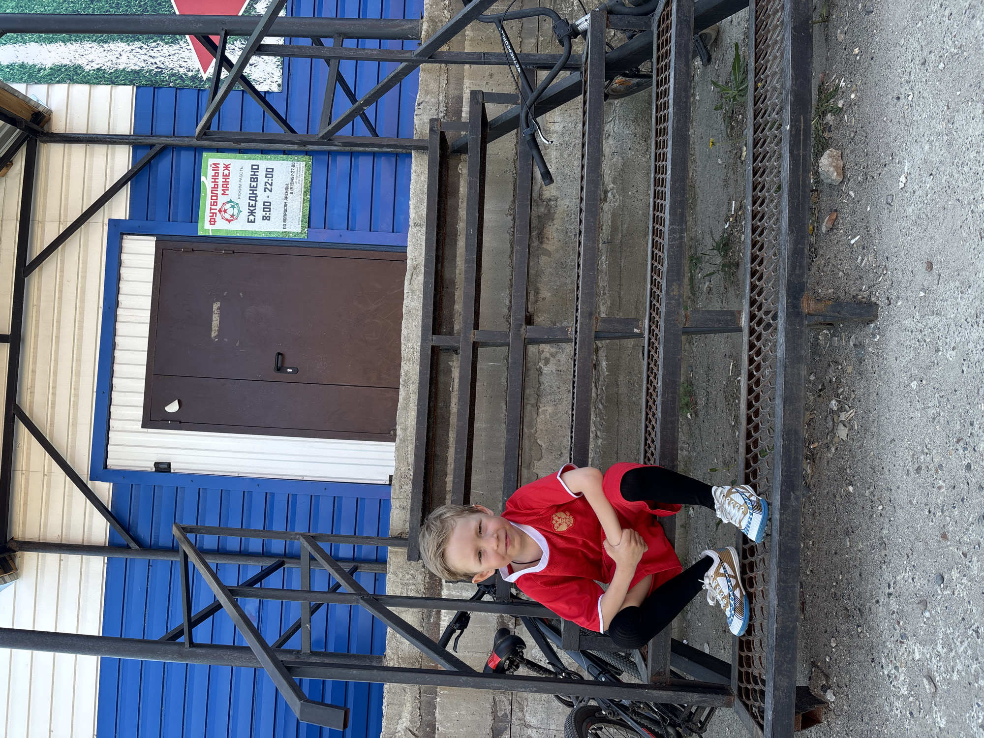 The photograph shows a young boy in a red soccer jersey with the flag of Russia on the chest. The boy is sitting on the metal steps of an outdoor staircase, with a black bicycle leaning against the staircase. The background features a building with blue and white corrugated metal siding and a dark brown door. Above the door is a sign in Russian, which translates to "Football Manege, Open Daily 8:00 - 22:00". The phone number 8 (919)457-27-80 is also displayed on the sign. The environment appears to be outdoors in Chaikovsky, Russia.
