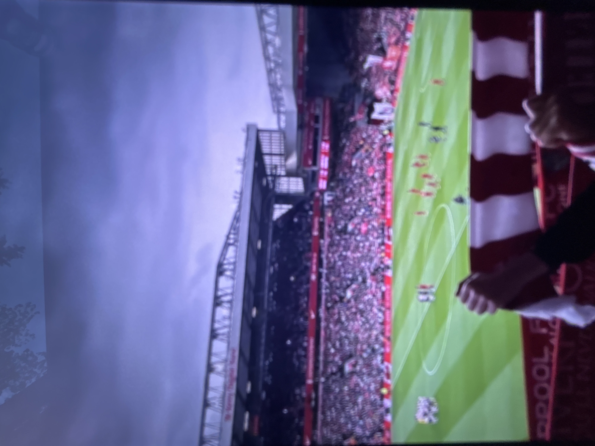 The image displays a photograph of a football (soccer) match being shown on a screen, with reflections and ambient light visible on the left side of the screen. The scene is a large, crowded football stadium, viewed from a high vantage point, looking across the pitch towards the opposite stands and the overcast sky.
A significant portion of the image is taken up by the stadium's architecture, including multi-tiered stands packed with spectators. The crowd is densely packed, with many individuals wearing red, suggesting support for the home team. Above the stadium, the sky is grey and cloudy, indicating an overcast daytime setting.
On the bright green football pitch, several players are visible. Some players are dressed in red kits, while others wear darker uniforms. White lines mark the playing area and sections of the field.
In the immediate foreground, to the right, a pair of hands holds up a prominent red and white striped banner or scarf. Visible text on this item clearly spells out "LIVERPOOL". Below this, further text is present but too blurred to decipher fully. This textual clue strongly identifies the team and suggests the location is Anfield stadium. The overall impression is that of an exciting and well-attended professional football match.